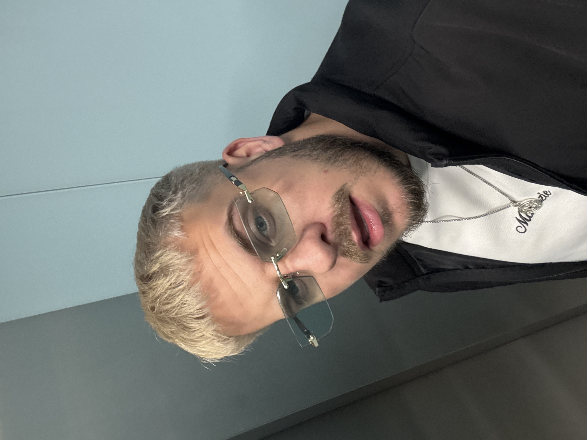 The image presents a close-up, rotated portrait of a young man, appearing as if the original photo was taken with the camera held vertically and then rotated 90 degrees counter-clockwise. He has short, light blond hair and a neatly trimmed blond beard. He wears rimless glasses with clear or very lightly tinted lenses. His blue eyes look directly at the viewer, and his tongue is slightly out, conveying a playful or cheeky expression.
He is dressed in a black jacket or hoodie layered over a white t-shirt. A silver chain with an intricate, circular pendant featuring a stylized design (possibly letters or an emblem) is visible around his neck. Part of a cursive word, "Moozie", is visible on his white t-shirt, beginning with "Mo...".
The background features a light blue wall on the left side of the frame, with a subtle horizontal line running across it. On the right, a darker gray surface, likely another wall or the floor, is visible. The scene appears to be indoors. The image captures a casual moment in Sofia, Bulgaria.