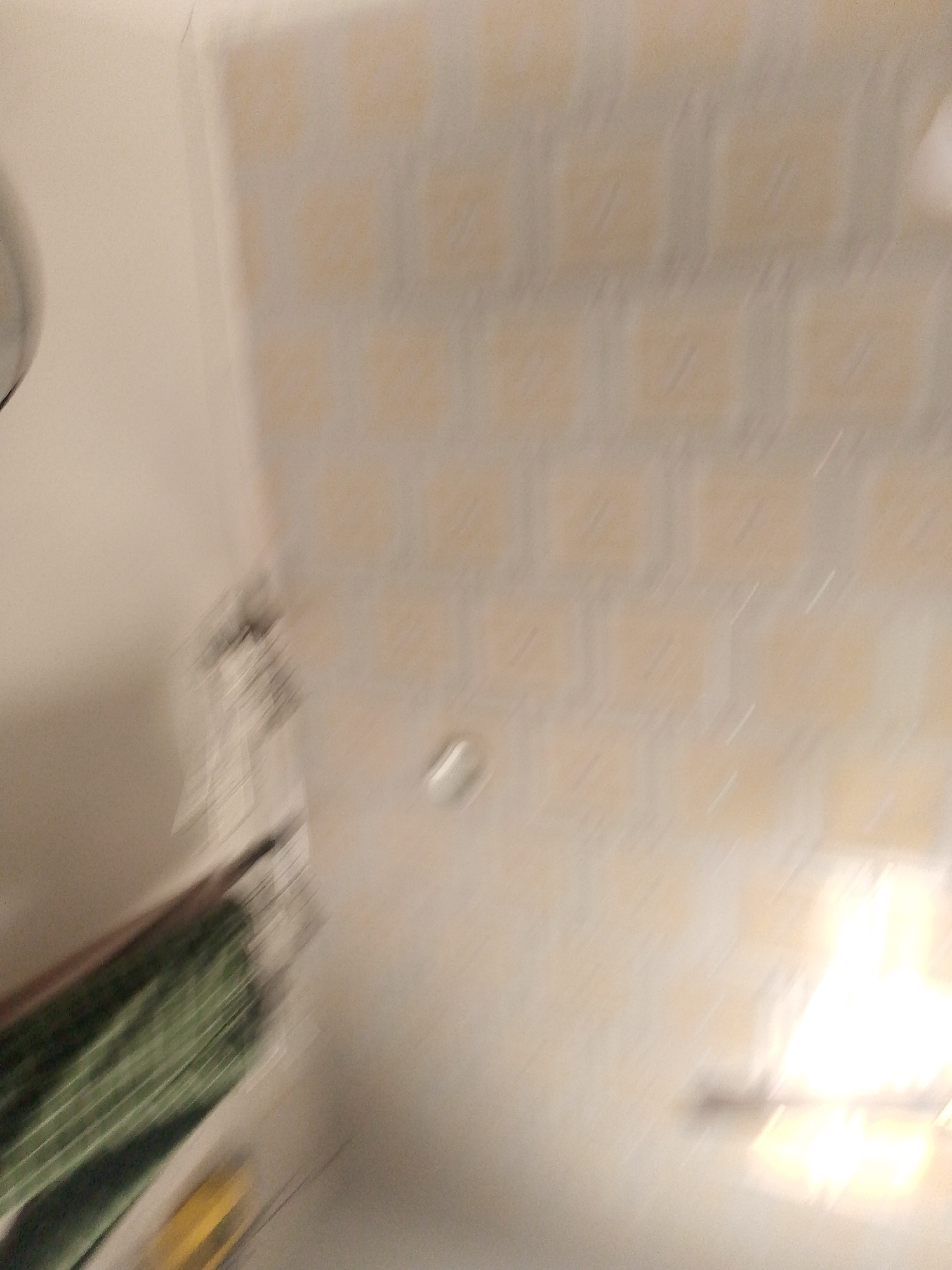 The image depicts an indoor scene, likely a room, with a blurry, abstract quality. The primary visual elements include a textured wall or ceiling with a repeating square pattern in pale yellow and white. In the lower left quadrant, a diagonal object with green and black stripes is visible, possibly a folded fabric or a piece of furniture. A bright, oblong yellow object is situated below this. The lighting suggests a light source in the upper right, creating a bright, blown-out area. There is a faint, circular object in the center of the frame. The overall impression is of movement or an out-of-focus capture. No specific location in Jauro Yinu, Nigeria, can be identified from this image due to its abstract nature and lack of discernible landmarks or contextual clues.