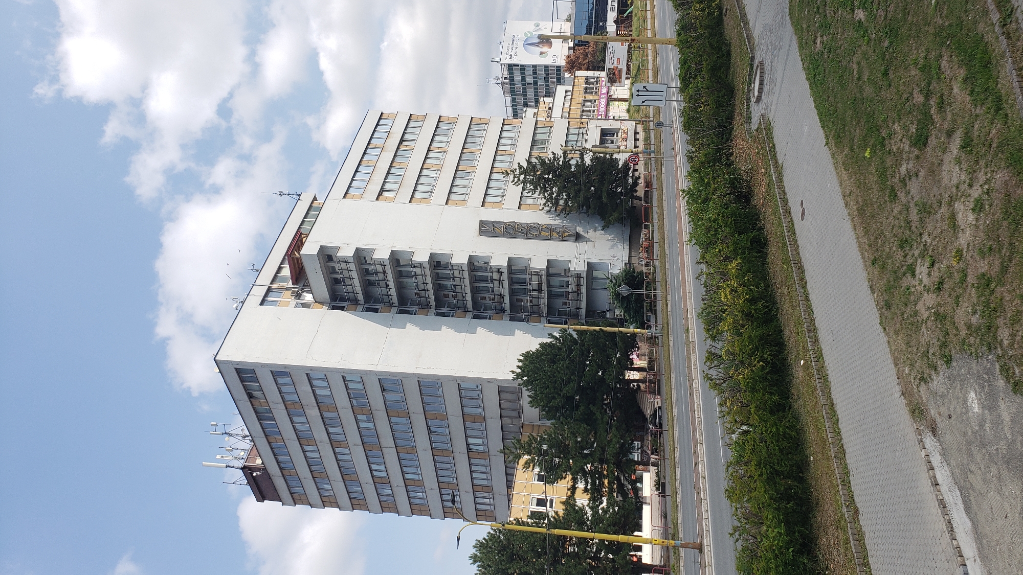 The image shows a tall, multi-story building with a predominantly white facade and many windows. The building is located in the city of Prešov, Slovakia, on a street with a sidewalk and a road. The building has a vertical sign on its side that appears to say "VzoroDEV". There are trees and bushes around the building, and the sky is visible with clouds. In the background, other buildings and structures can be seen, including a taller, darker building with advertisements on its side. The scene appears to be during the daytime, with clear weather. There are no people visible in the image.