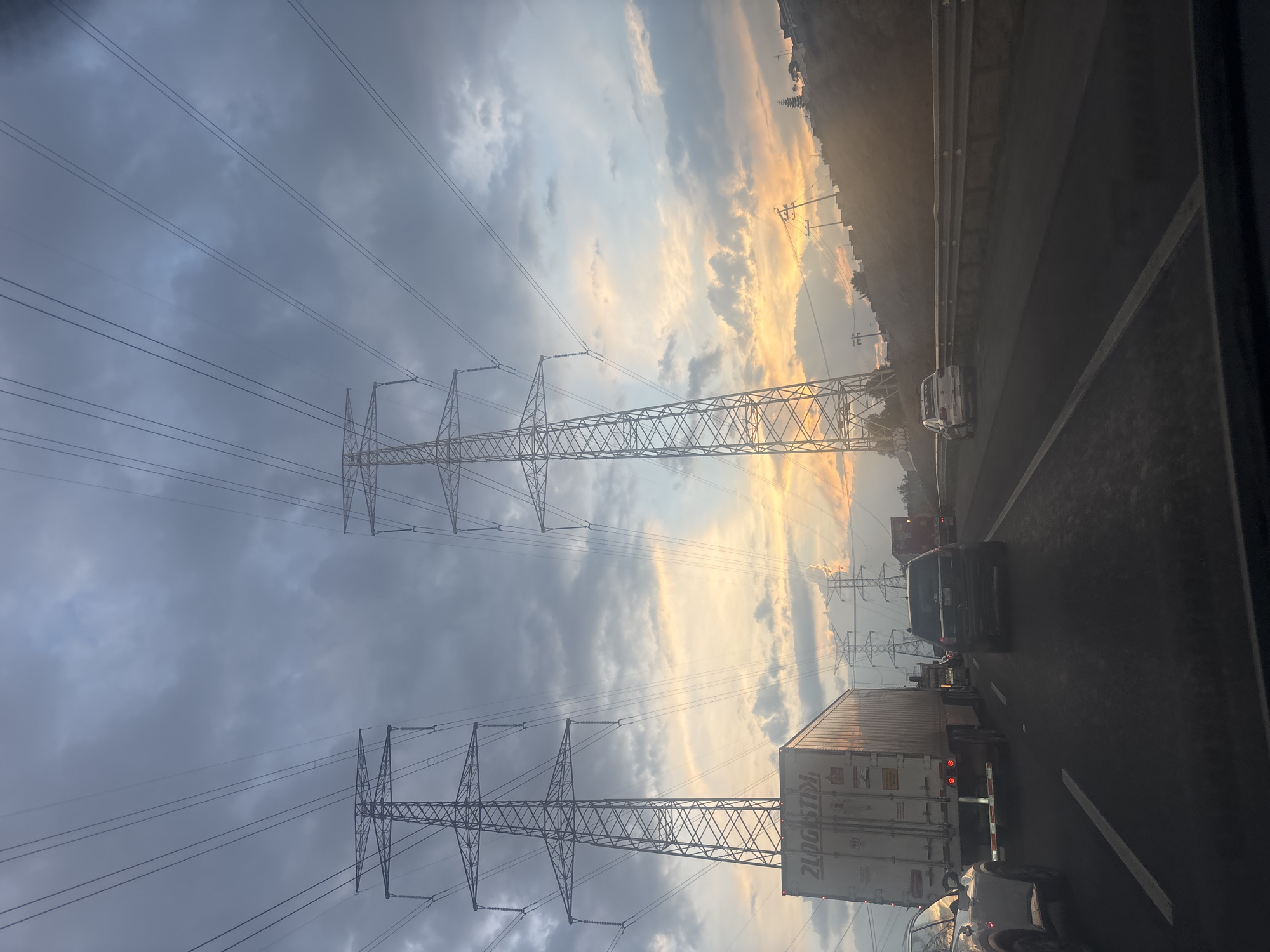 The image depicts a traffic jam on a highway near Cuautitlán Izcalli, Mexico, at sunset. Two metal power line towers are visible against a cloudy sky with patches of orange sunlight. Cars and trucks are lined up on the road, including a large white semi-trailer truck. A guardrail is visible along the side of the highway. The image appears to have been taken from inside a vehicle.