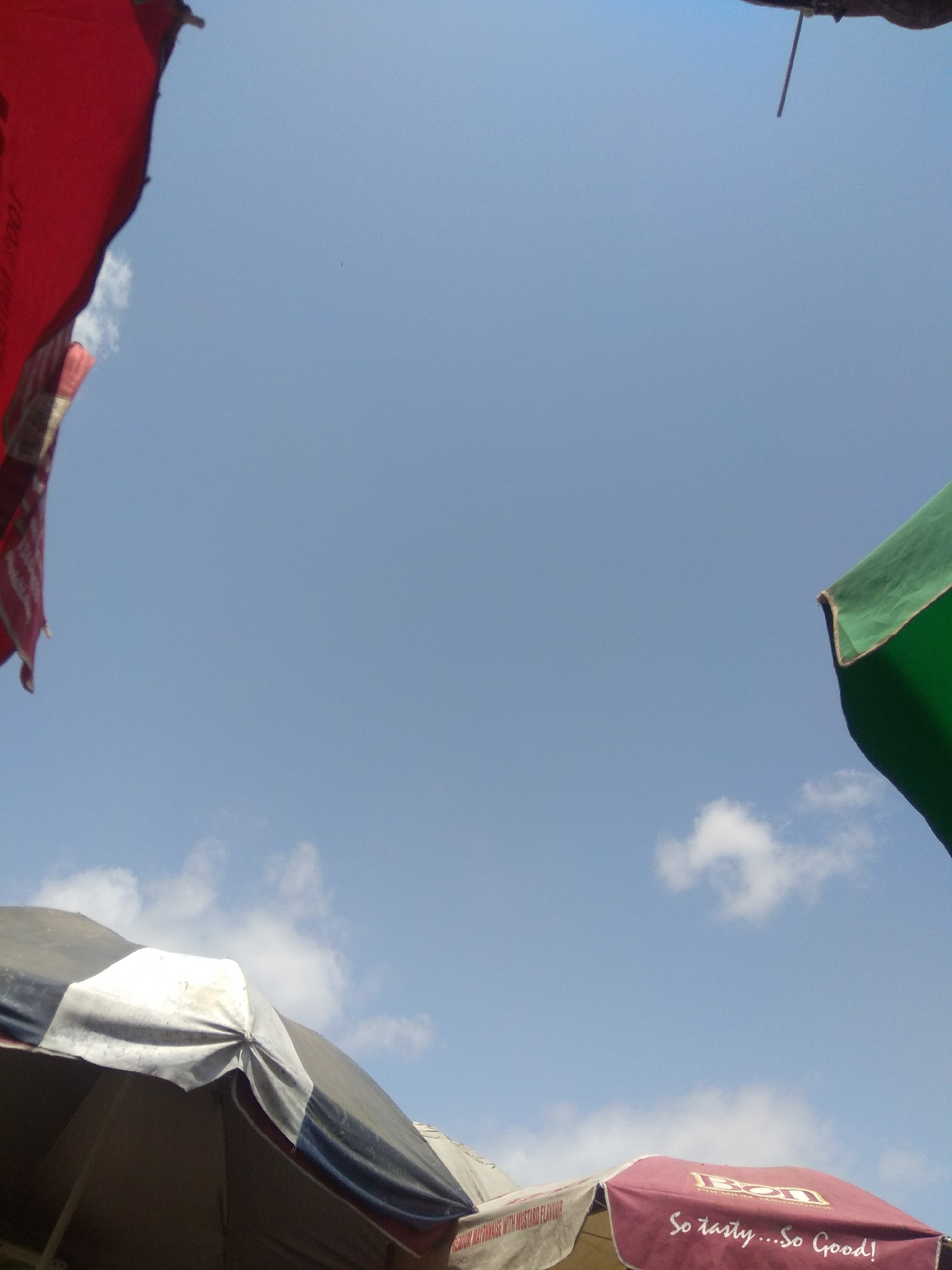 In the city of Jalingo, Nigeria, a low-angle outdoor shot captures a sky with scattered white clouds against a pale blue backdrop. Three umbrellas are visible, suggesting a marketplace or outdoor gathering. On the left, a portion of a large red umbrella with faded white text is prominent. In the upper right quadrant, the edge of a green umbrella is visible. In the lower part of the frame, a striped umbrella with black, white, and grey sections is angled upwards, revealing its structure. To its right, a maroon umbrella displays the brand "Bonn" and the tagline "So tasty...So Good!" in white script. The scene appears to be during daylight, with the sun likely overhead or slightly behind the camera, casting no distinct shadows. No people are directly visible, but the presence of umbrellas implies human activity.