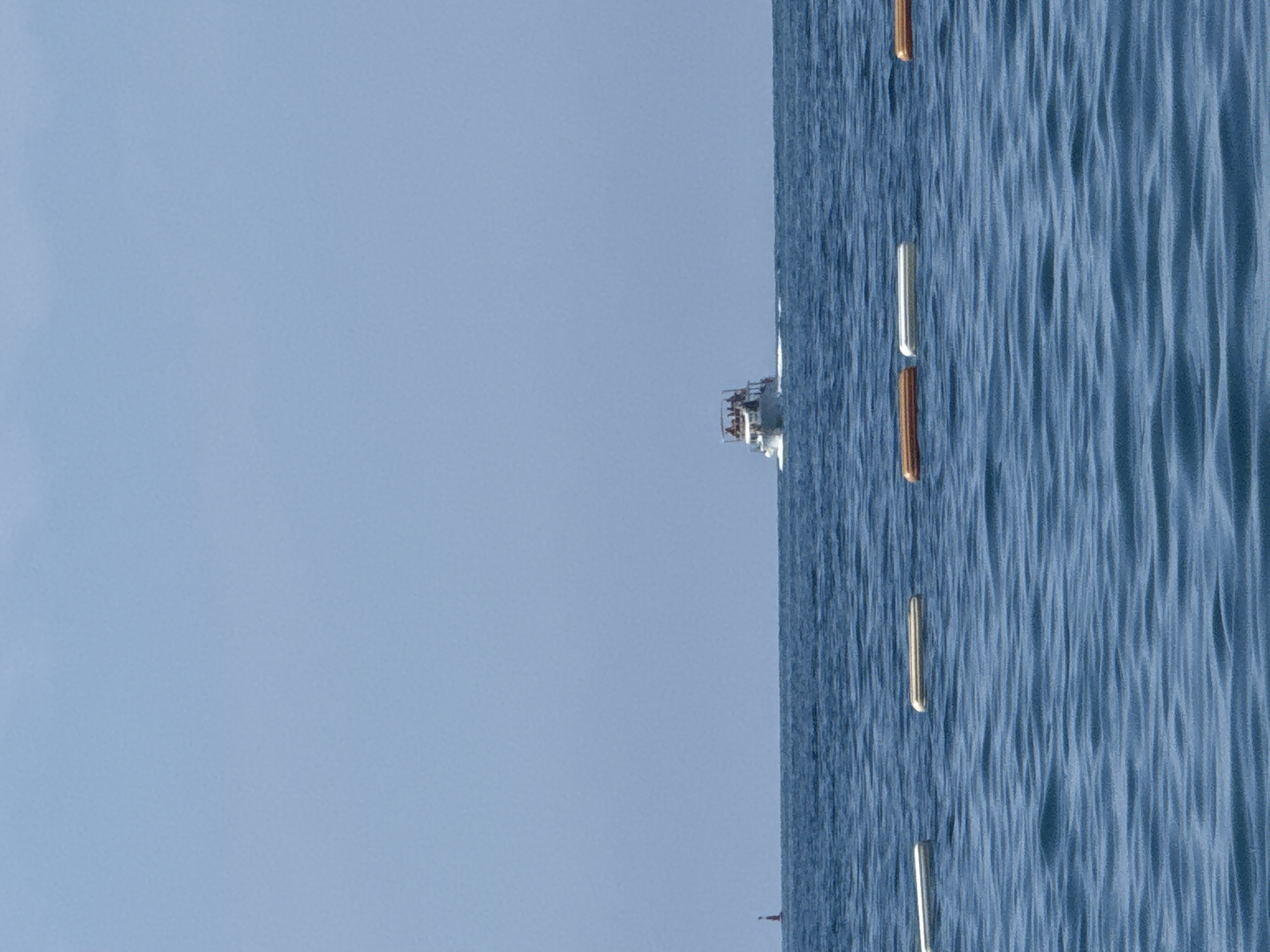 The image depicts a tranquil scene on the water. In the distance, a white boat with several people on board cruises across the blue expanse, leaving a white wake behind it. The horizon is clear, with a pale blue sky overhead. Closer to the viewer, a line of buoys, some white and some orange, floats on the surface of the gently rippling water, marking a boundary or a designated area. The overall environment suggests a calm day at sea, possibly near the coast. No specific landmarks are identifiable to pinpoint the location within Boreti, Montenegro, but the setting is consistent with a coastal area.