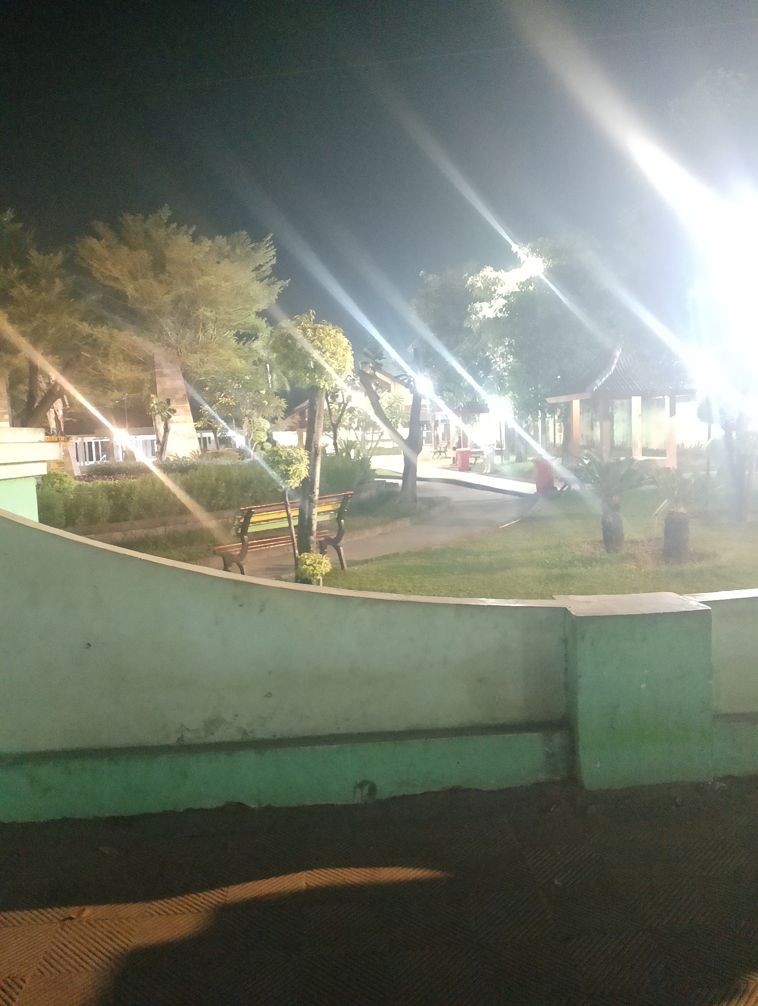 The image captures a park at night, illuminated by bright artificial lights that create lens flares and streaks. In the foreground, a light green wall runs horizontally, with a darker textured surface below it. Beyond the wall, a grassy area is visible, with a park bench featuring a wooden seat and a green and yellow striped back. Further in the background, trees with sparse foliage are present, and a small, open-sided pavilion with a dark, sloped roof stands amidst the greenery. The overall environment suggests a public outdoor space, likely a recreational park in the city of Ngadiluwih, Indonesia, given the context. There are no discernible people or specific actions taking place in the scene.