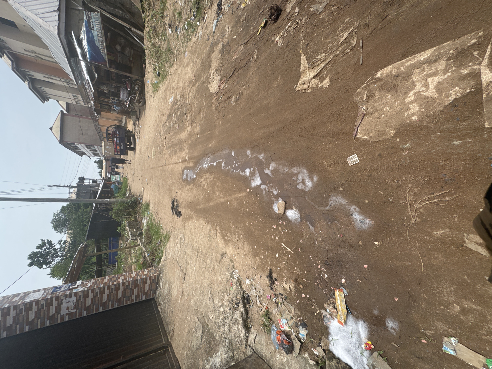 The image depicts a dirt road in Ihiagwa, Nigeria. Along the left side are buildings, one appearing to be a shop. The road is littered with debris, including plastic, paper, and what appears to be refuse. Patches of white substance, possibly detergent or a similar material, are visible on the road surface. In the distance, figures of people and vehicles are present. The general environment appears unpaved and urban, with signs of human activity and waste.