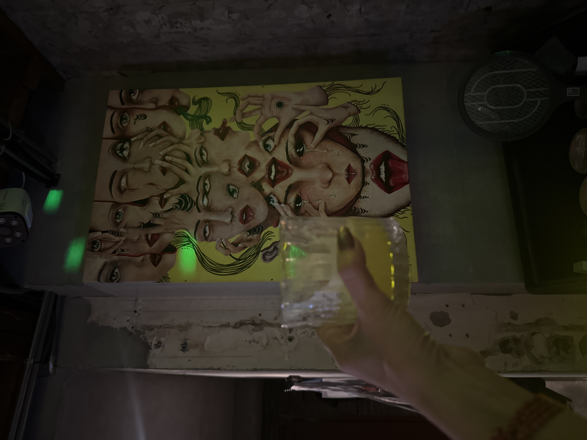 The image depicts an indoor scene. A hand holds a glass of a yellow liquid. On the wall behind the hand, there is a painting of faces. To the right of the painting is a black electric fly swatter. A small light fixture is visible on the wall to the left. The wall is painted in a dark gray color and shows signs of aging and wear. The setting appears to be a bar in Xuhui District, China.