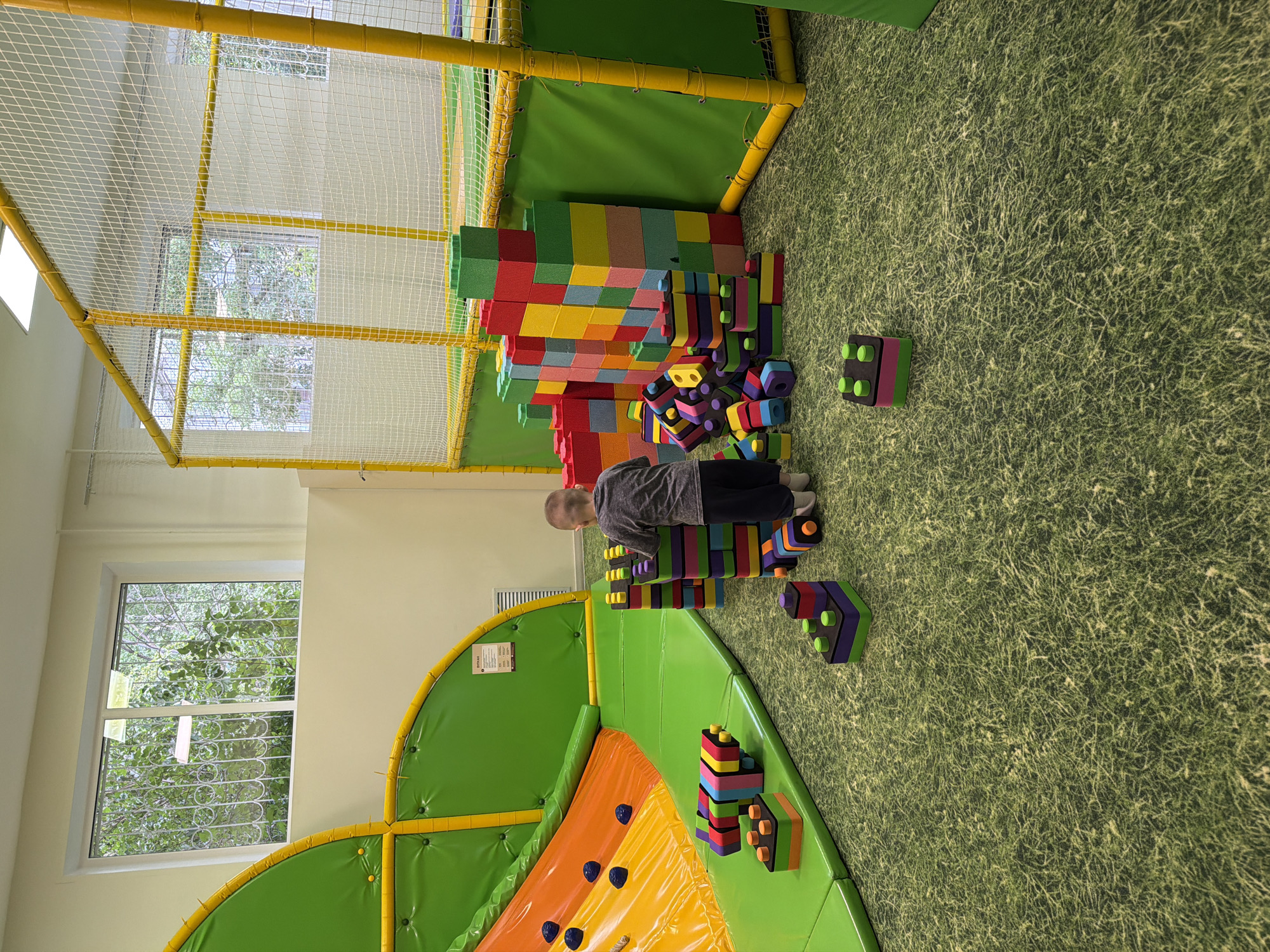 A young boy plays with large, colorful foam blocks in an indoor playground. The playground is located in Chaikovsky, Russia. The boy is building a structure with the blocks. The floor of the playground is covered in a green, textured surface resembling grass. There are other play structures nearby, including a climbing wall and a net enclosure. Sunlight streams in through a window.