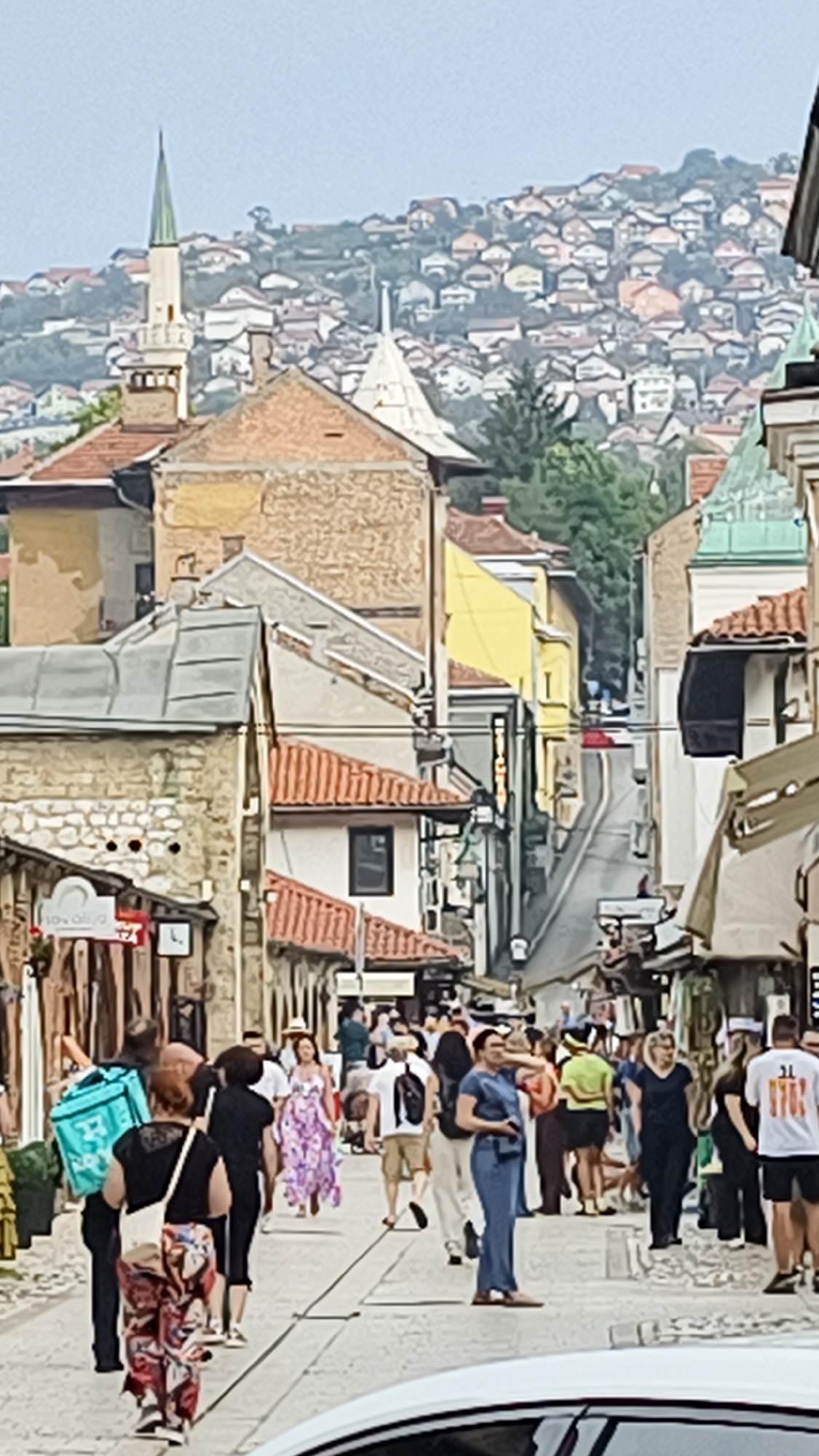 The image shows a street scene in Sarajevo, Bosnia and Herzegovina. In the background, a hillside is covered with densely packed houses, characteristic of the city's urban sprawl. A minaret of a mosque rises from the left side of the image, with a green spire, hinting at the religious and architectural landscape of Sarajevo. The foreground depicts a bustling street lined with buildings, some with red tile roofs. People are walking along the cobblestone street, engaged in various activities. One person is carrying a large turquoise cooler bag. Another person in a long floral dress is walking towards the camera. Shops and stalls appear to be present along the sides of the street, with awnings providing shade. The overall impression is one of a lively urban environment, possibly a market or shopping district, with a distinct cultural and historical ambiance. The lighting suggests it is daytime, possibly late morning or afternoon.