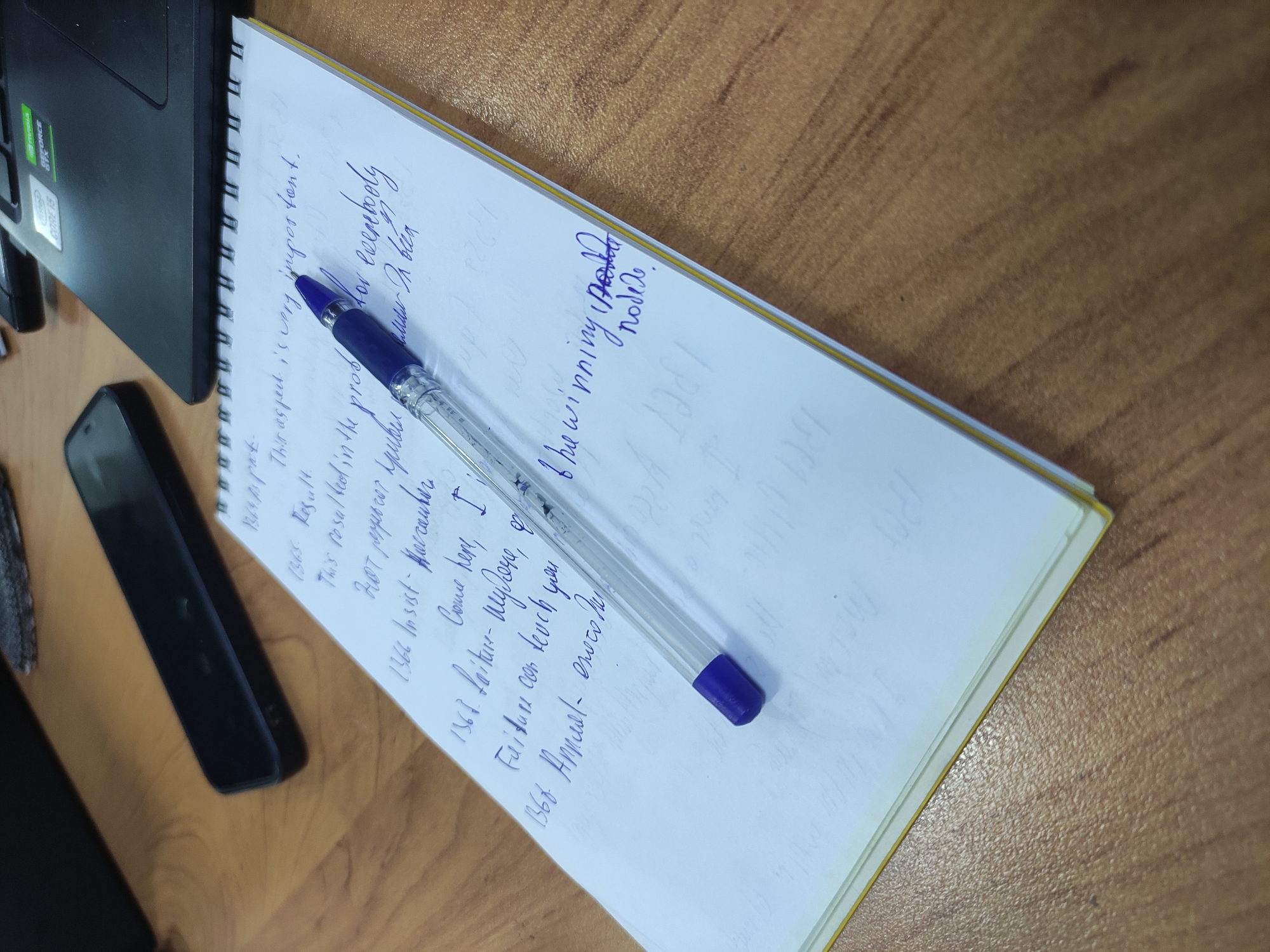 An overhead, close-up shot shows a notebook with blue ink handwriting on a wooden desk. The notebook is open to a page with several numbered entries, the top of which reads "1364. Part". Below that, "This aspect is very important." A pen with a blue cap lies across the notebook, pointing towards the right. Further down the page, entries include "1365. Result. This result led in the problem", "1367. paper car spider - for everybody", "1366. Insist - Harranhur", "Come here, I", "Faidure can levch you", and "1366 Annual- eroco ho". At the bottom right of the notebook page, a scribbled phrase reads "The winning 1221 node". To the left of the notebook is a black smartphone, and to the right, the corner of a black laptop is visible. The wooden desk has a warm, brown, textured surface.