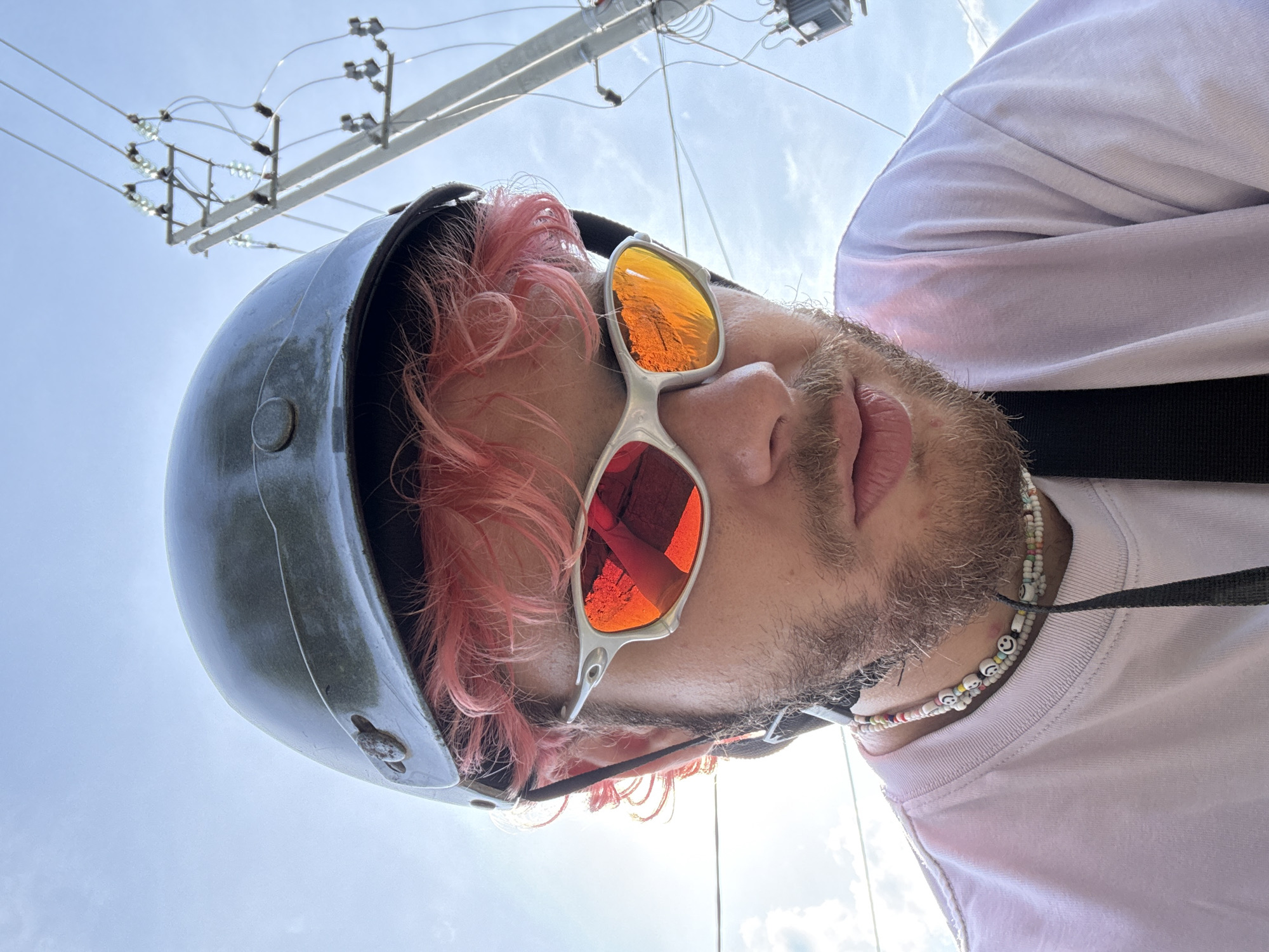 A low-angle shot depicts a person wearing a gray helmet, orange/red reflective sunglasses, and a light-colored t-shirt. The person has pink hair and a beard. A beaded necklace is visible around their neck. Overhead, power lines and utility poles are visible against a blue sky. The reflection in the sunglasses suggests a cityscape. While the prompt requests the location of Đà Nẵng, Vietnam, this cannot be determined from the photograph alone.