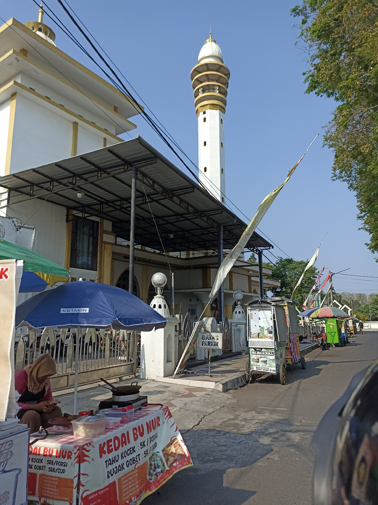 The image depicts the exterior of a mosque in Lumajang, Indonesia, bathed in bright daylight under a clear blue sky. A prominent white minaret with golden accents stands tall, while the main mosque building, also white with yellow trim, features a covered walkway extending from its facade. In front of the mosque, several street vendors operate their stalls. One vendor, a woman wearing a light brown hijab and an apron, is seated at a table laden with food supplies, preparing to cook. Her stall displays a banner with the text "KEDAI BU NUR" and prices for various food items like "Tahu Kocok" and "Rujak Gobet." Adjacent to her, a small cart with the label "BURGERKU" and a phone number suggests another food vendor. Further down the street, additional colorful stalls and small vehicles indicate a lively market scene. The presence of electrical wires overhead and trees lining the street provides context to the urban environment. The overall impression is that of a typical day in a bustling area near a religious institution in Lumajang.