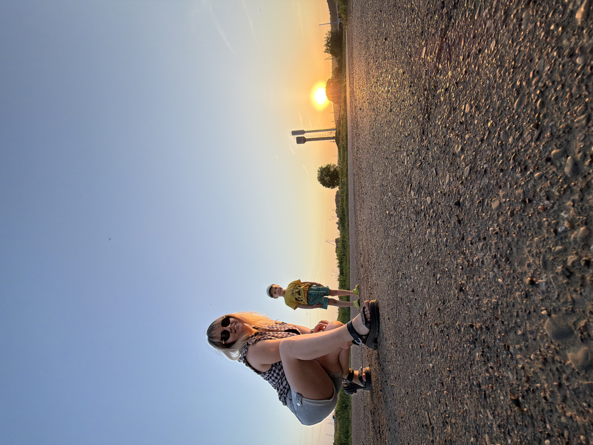 A low-angle photograph shows a woman and a child standing on an asphalt road at sunset. The woman is crouching and facing the camera, wearing sunglasses, a checkered shirt, and denim shorts. She is wearing black sandals. Behind her, a young boy stands, wearing a yellow t-shirt, blue patterned shorts, and a cap. In the background, the setting sun shines between two tower-like structures. A small tree is visible to the right. The location is likely in the city of Чумна, Russia, based on the user's prompt.