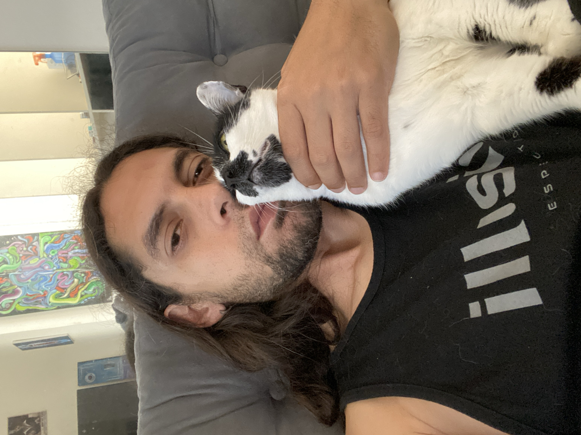 An indoor, close-up portrait shows a man and a black and white cat cuddling on what appears to be a grey upholstered couch or bed. The image is oriented with the man lying horizontally, his head resting on the grey surface. He has long, dark hair, a beard, and dark eyes, looking directly towards the camera. He is wearing a black tank top.
The cat, primarily white with black markings around its face, ears, and body, is nestled against the man's face. One of the man's hands is gently resting on the cat's back, holding it close. The cat's face is pressed against the man's cheek, showing a moment of affection between the two. The cat has yellowish-green eyes.
In the background, behind the man's head, parts of a room are visible. There are light-colored walls and several pieces of art, including a vibrant, abstract, multi-colored painting hung horizontally. Below it, a reflection or another artwork shows what looks like a blue object and some white shelves with bottles.
Visible text on the man's black tank top includes partial letters that spell out "ILLUSION" or similar, and "ESPACIO" or "ESPECIO." The scene suggests a relaxed and affectionate interaction, likely during the daytime, in a home in Buenos Aires, Argentina.