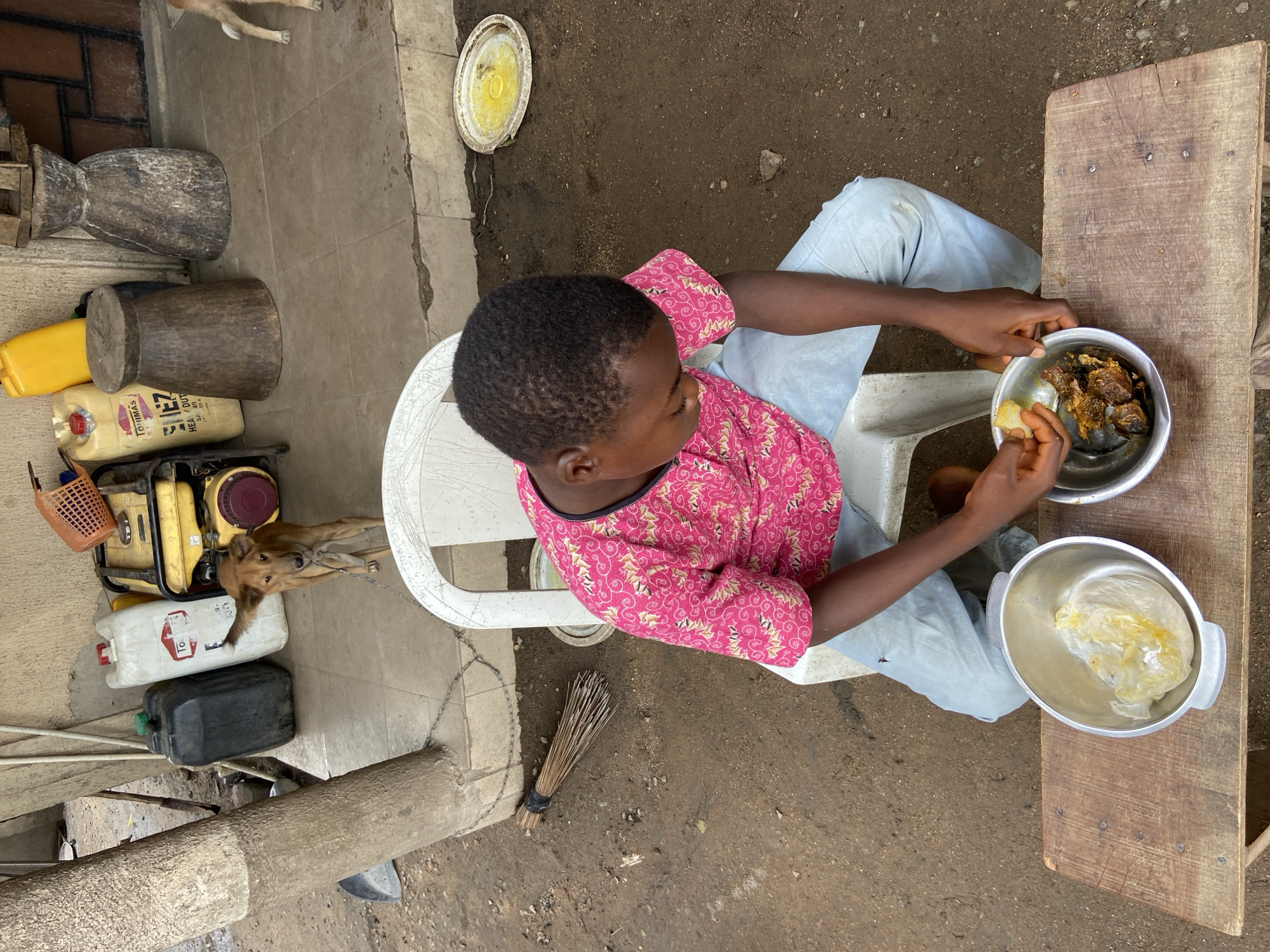 An overhead view shows a young person seated on a white plastic chair eating from a silver bowl while positioned at a wooden table. The person is wearing a pink patterned shirt and light blue pants. On the table next to the bowl of food is another silver bowl. The area surrounding the person is a mix of dirt and concrete. A broom lies on the ground next to the chair. A dog, tethered by a chain, stands near a portable generator, jerry cans, and wooden vessels. The location is Mararaba, Nigeria.
