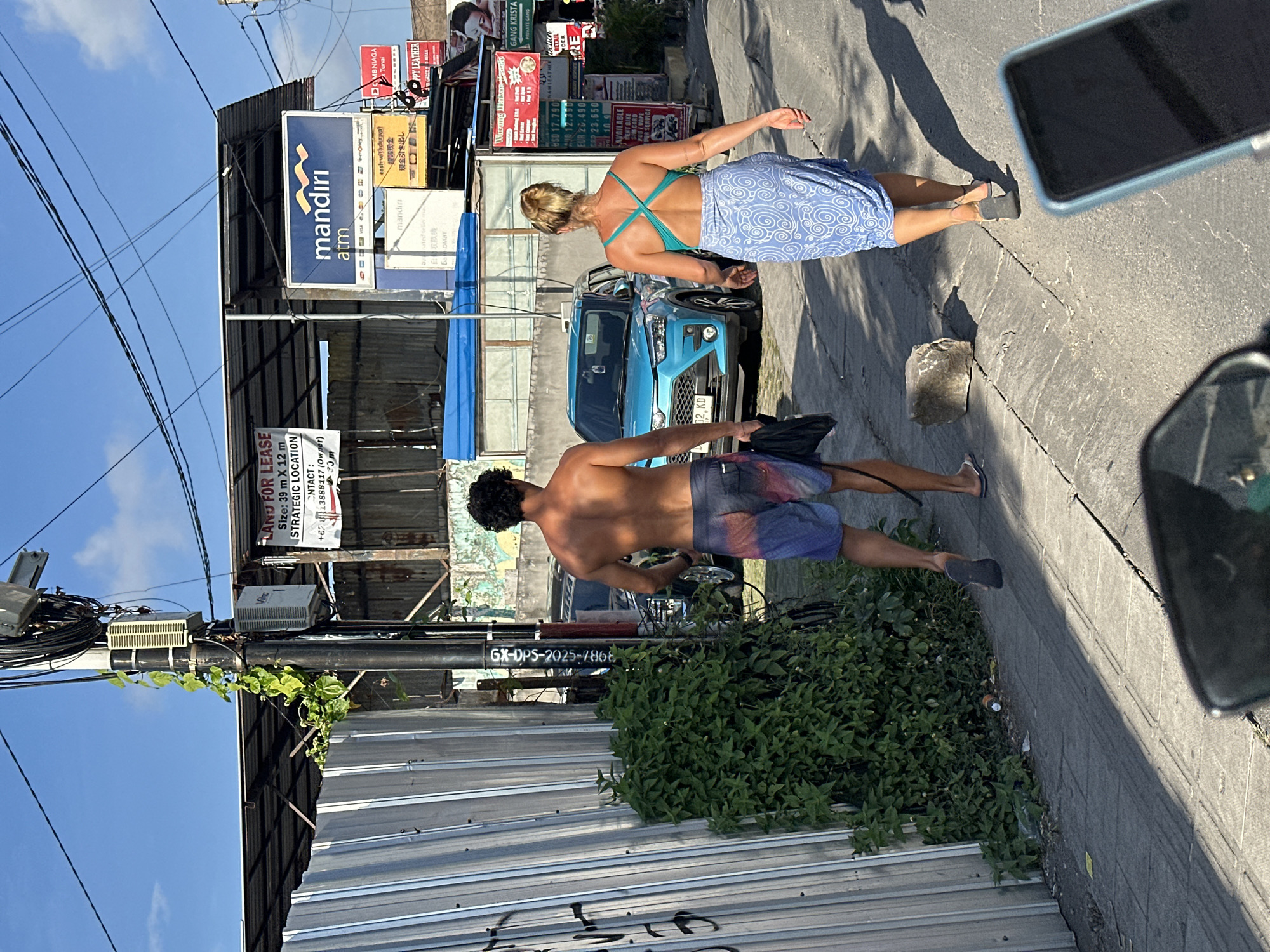 The image captures a street scene in North Kuta, Indonesia, under a clear blue sky with scattered clouds. In the foreground, the rear view of a man and a woman walking away from the camera is visible. The man, shirtless, wears multi-colored board shorts and flip-flops, carrying a black bag in his right hand. The woman is dressed in a turquoise halter-neck top and a blue sarong with a white swirl pattern, also wearing flip-flops.
A bright blue SUV is parked on the right side of the road, and a metal fence runs along the left side of the image, with a dense green bush growing next to it. A utility pole with numerous cables and a small electrical box is prominently featured on the left. Signs are visible on buildings in the background, including one for "mandiri atm" and another advertising "LAND FOR LEASE." The environment appears to be an urban or semi-urban area with paved roads and sidewalks. The lighting suggests it is daytime, likely late afternoon, given the shadows cast on the ground.