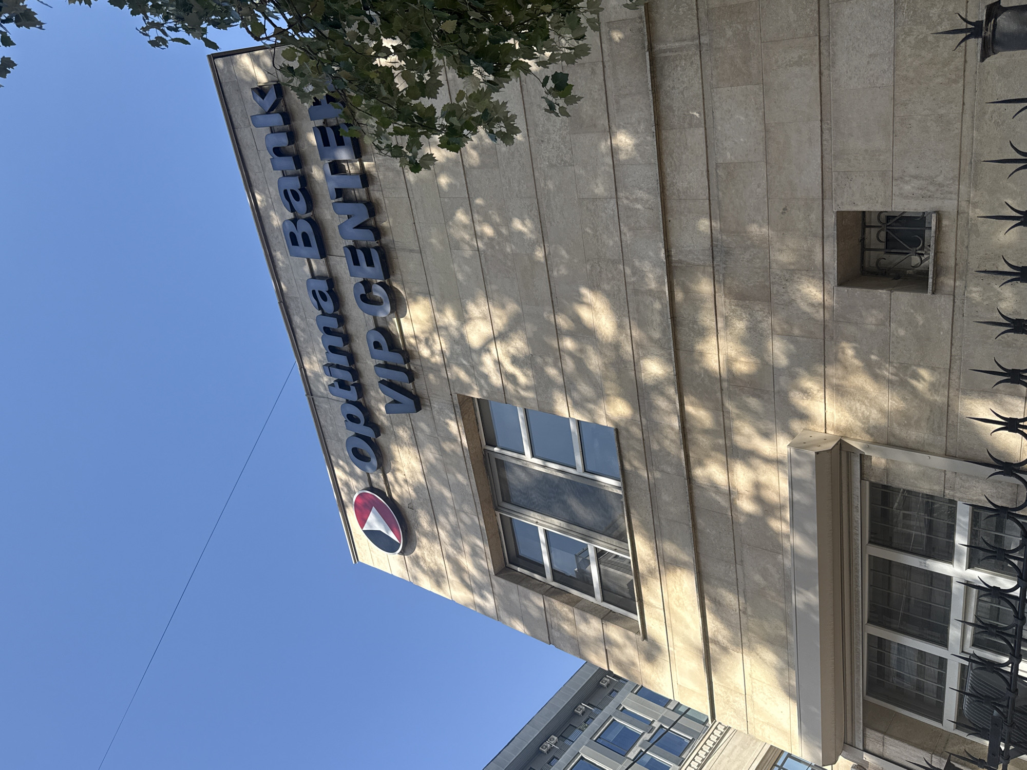 A low-angle outdoor shot captures the facade of a building in Bishkek, Kyrgyzstan, under a clear blue sky. The building, constructed with light-colored stone blocks, displays signage for "Optima Bank VIP CENTER" in dark blue lettering. A circular logo featuring a red and white design is positioned to the left of the bank's name. The building has multiple windows, some with reflective glass. The facade is partially obscured by shadows cast by trees, with some tree branches and leaves visible in the upper right corner. A black wrought-iron fence runs along the bottom of the image. To the left, the corner of another building with multiple windows is visible. The overall scene is devoid of people and depicts a commercial establishment.