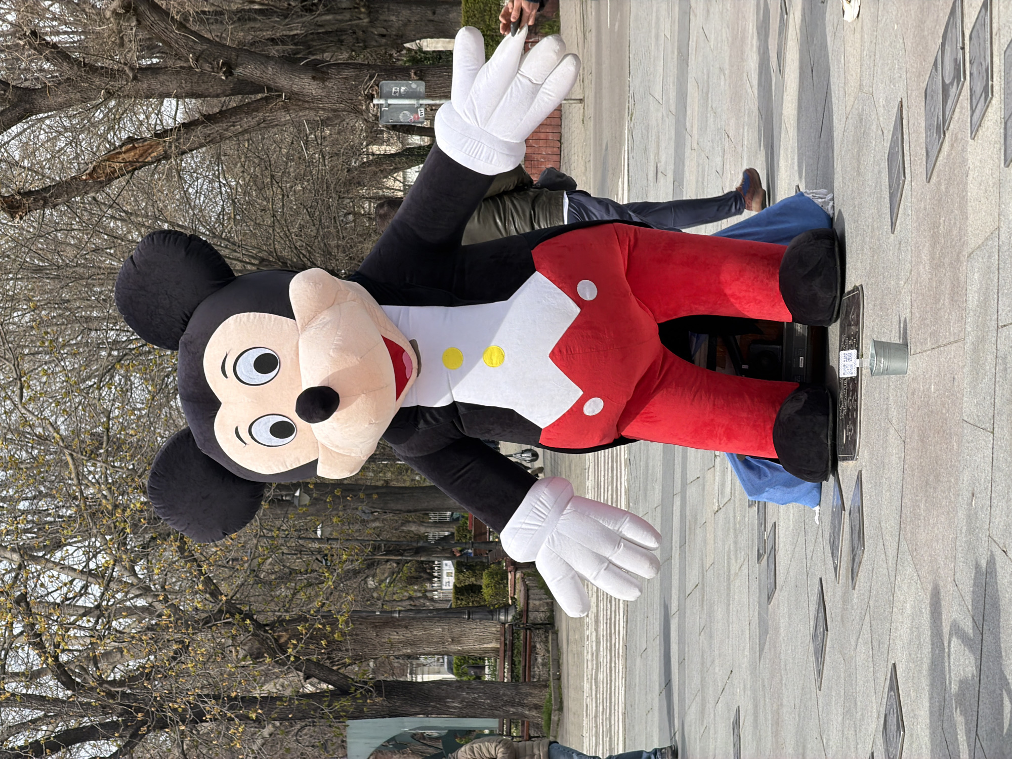 A large, plush Mickey Mouse figure lies on its side on a paved surface. The figure is wearing white gloves, a black jacket, and red pants. The setting appears to be an outdoor public space, possibly a pedestrian zone. A few people are standing nearby. The background includes trees and what may be street furniture. The surface of the pavement is adorned with metal plaques of names, possibly the Walk of Fame in Varna, Bulgaria.
