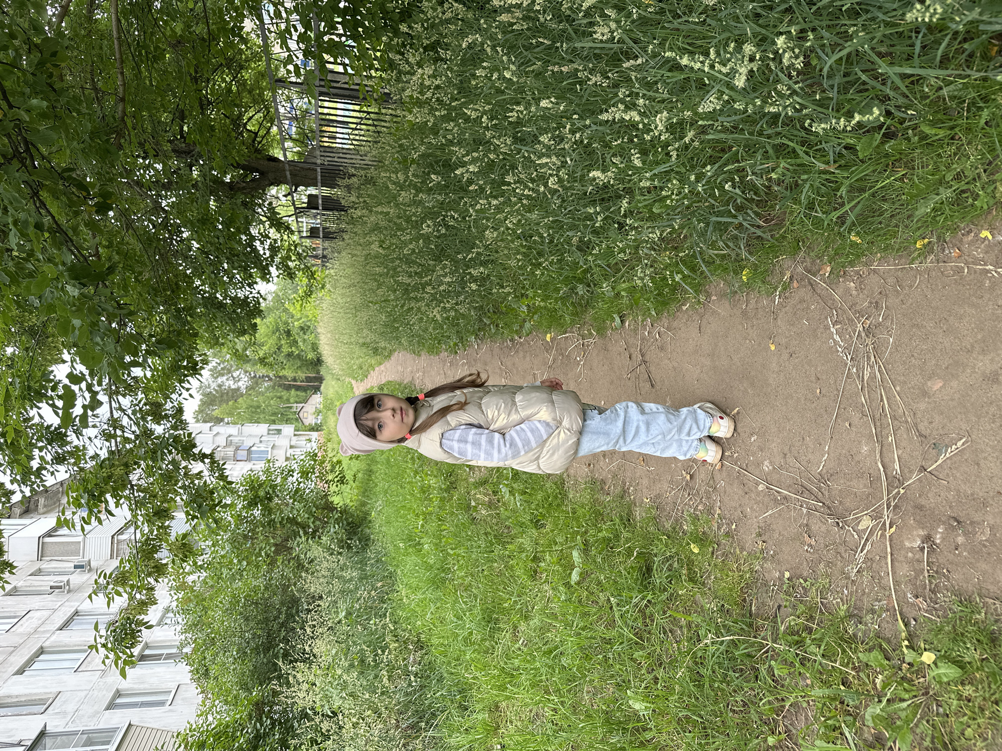A young girl stands on a dirt path lined with tall grass and trees, in Chaikovsky, Russia. The girl, wearing a light-colored puffer vest, striped shirt, and light blue jeans, faces the camera with her hands in her vest pockets. She has long brown hair pulled into two pigtails and wears a light-colored beanie with ear details. Behind her, a fence separates the path from a patch of tall grass with white flowers, and further back, a large apartment building is visible. The overall scene depicts an outdoor setting on a cloudy day, likely in a residential area.