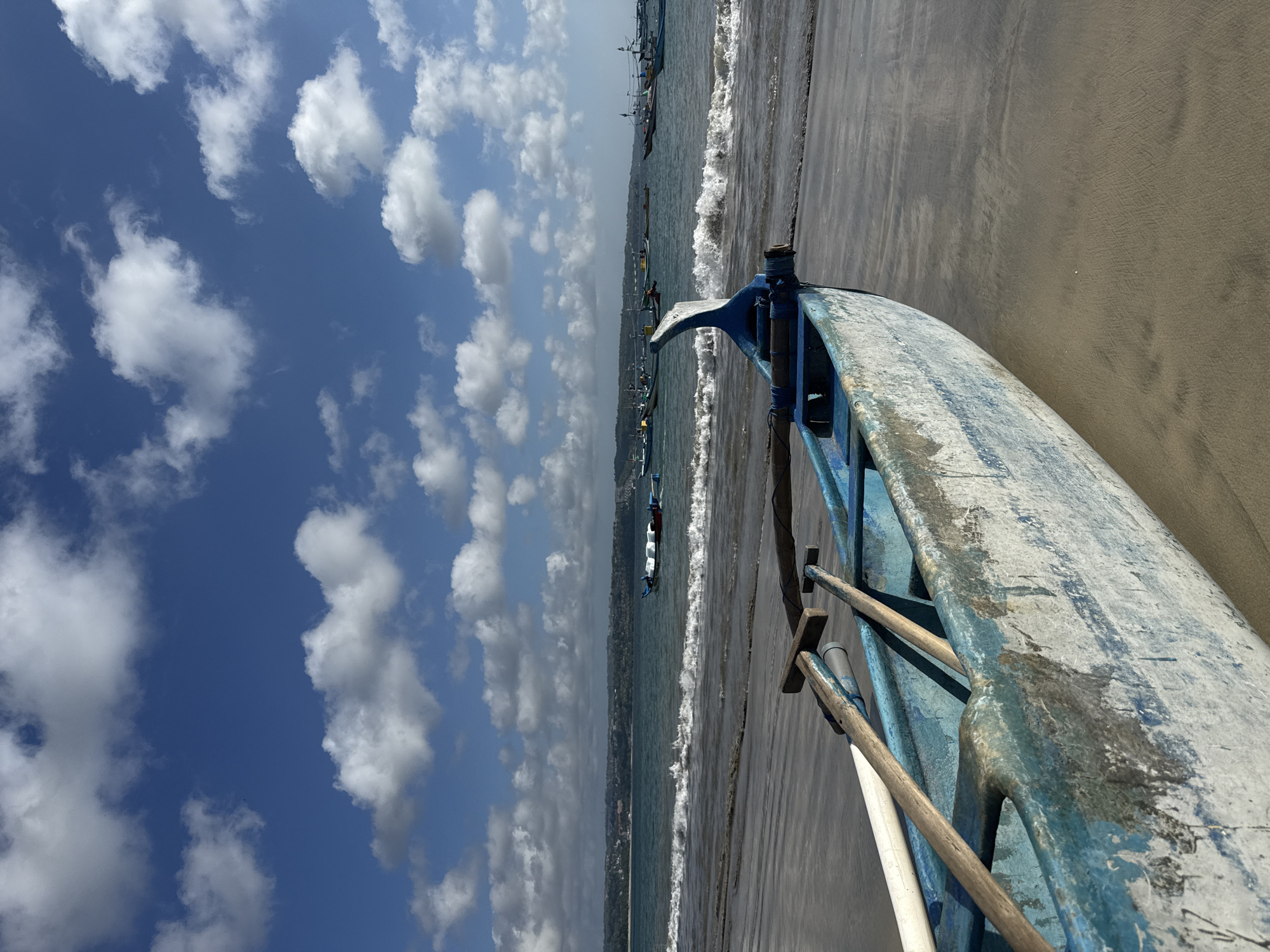 The image shows a view of Kuta Beach in Bali, Indonesia, from the perspective of a boat pulled up on the sand. The boat is painted with blue trim and white paint. The sand is coarse and dark. The sea is slightly turbulent with small waves. The sky is blue, with scattered cumulus clouds. A few boats are seen out in the water, and the coastline of Kuta is visible in the background.