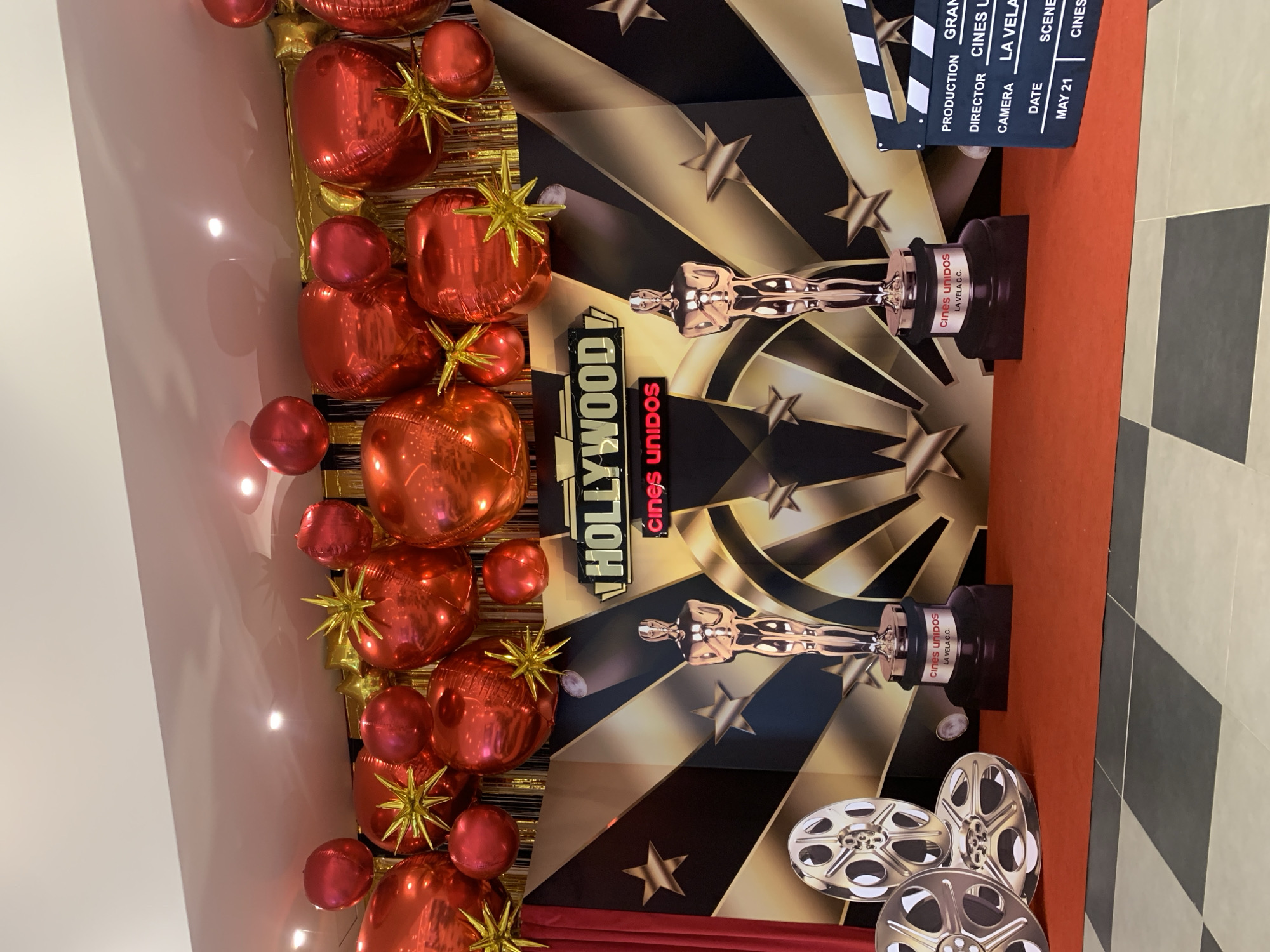 Here is a description of the image:
This image captures a movie premiere event or celebration, potentially at the Cines Unidos cinema complex in La Vela C.C., Porlamar, Venezuela. The setting is indoors, with a backdrop adorned with cinematic elements. The background features a poster design with gold and black starburst patterns, depictions of an Oscar-like statuette, and the words "HOLLYWOOD" and "cines UNIDOS." A film clapperboard graphic with the words "PRODUCTION GRAM," "DIRECTOR CINES U," "CAMERA LA VELA," "DATE," "SCENE," and "MAY 21" is also visible. A garland of shiny red balloons, some decorated with golden star-shaped embellishments, decorates the left side of the frame. Simulated film reels sit on the floor toward the lower part of the image. The floor is a black and white checkered pattern.