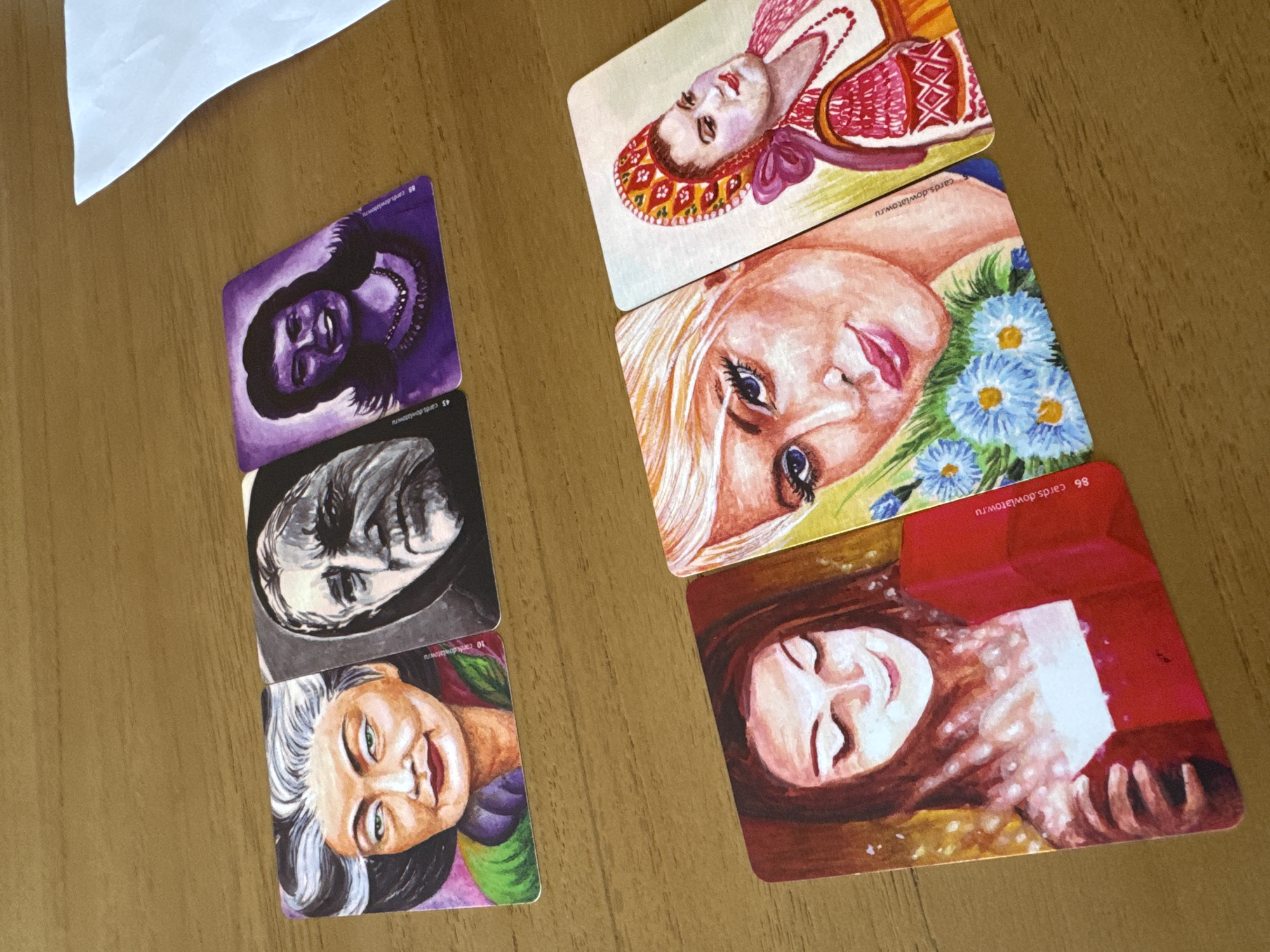 The image displays six vividly illustrated cards, likely oracle or tarot, laid out on a light brown wooden surface. They are arranged in two vertical stacks of three. A white piece of paper is partially visible in the top-left corner.
The leftmost stack, from top to bottom, shows:
1. A purple card featuring a smiling woman with dark skin, dark hair, and a necklace. The text "89 cards.dowlatow.ru" is visible.
2. A dark-toned card depicting a person's face with a contemplative or solemn expression, rendered in black and white or grayscale. The text "45 cards.dowlatow.ru" is visible.
3. A colorful card with an older woman sporting grey hair, green eyes, and a warm smile, dressed in purple and green. The text "10 cards.dowlatow.ru" is visible.
The rightmost stack, from top to bottom, shows:
1. A card featuring a woman with a traditional-looking red and gold patterned headdress, light skin, and red lips.
2. A card depicting a young blonde woman with bright blue eyes, looking upwards, surrounded by blue and white flowers. The text "5 cards.dowlatow.ru" is partially visible.
3. A red-backed card showing a brown-haired woman with closed eyes and a soft smile, appearing to interact with a glowing object amidst sparkles. The text "86 cards.dowlatow.ru" is visible.
All card illustrations are in a distinct watercolor or painted style, focusing on facial expressions and portraits that convey a range of emotions from joy and serenity to contemplation. The cards are neatly arranged, suggesting a display, reading, or preparation. The bright, even lighting indicates a daytime setting, likely indoors on a wooden table.