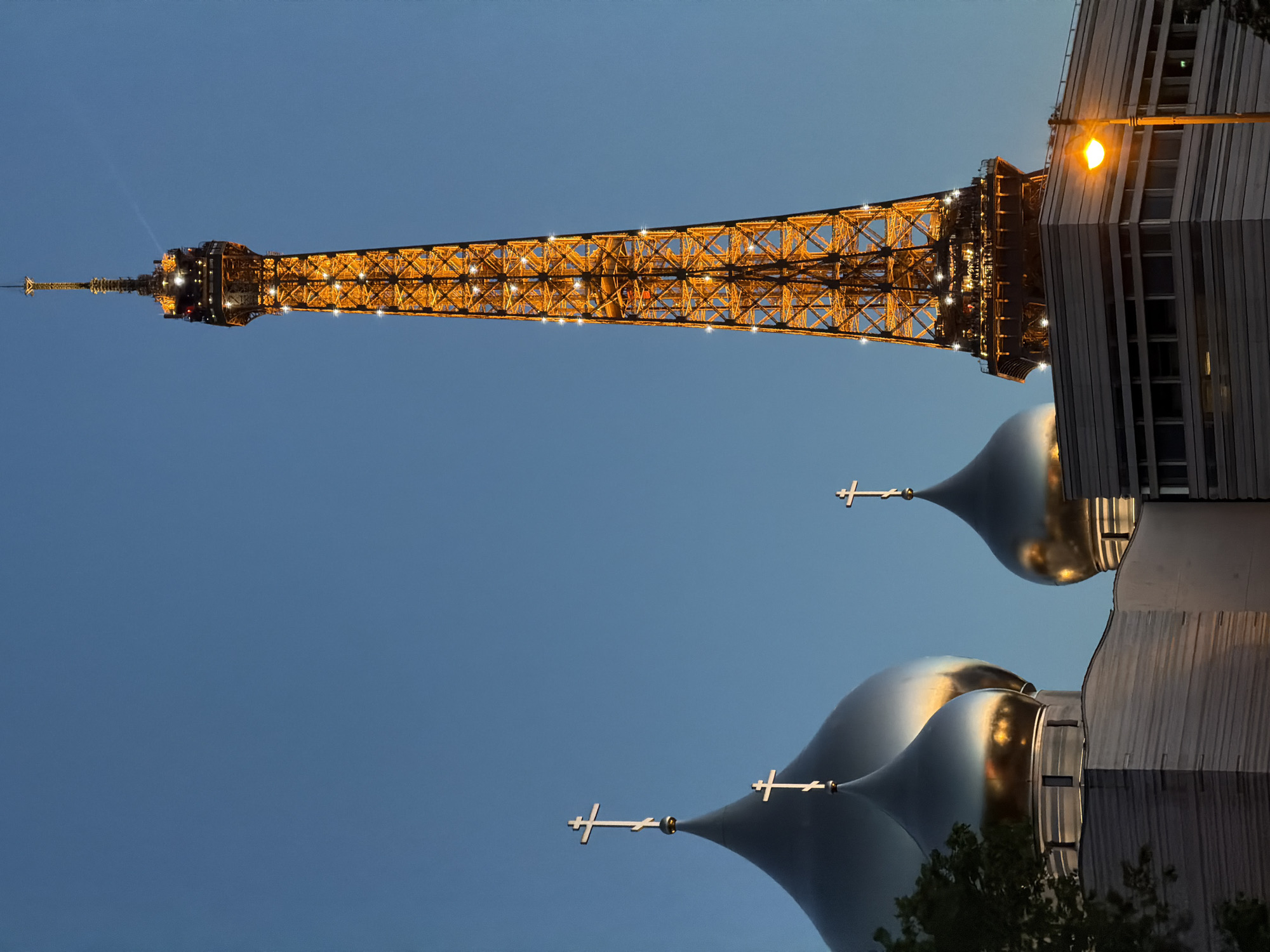 The image features the Eiffel Tower in Paris, France, set against a clear blue sky. The tower is illuminated with golden lights, highlighting its intricate metal framework. In the foreground, the silver domes of the Holy Trinity Cathedral stand out. The cathedral's domes are topped with golden crosses. A building with a modern facade, lit by a street lamp, appears next to the cathedral. The image is oriented upside down.