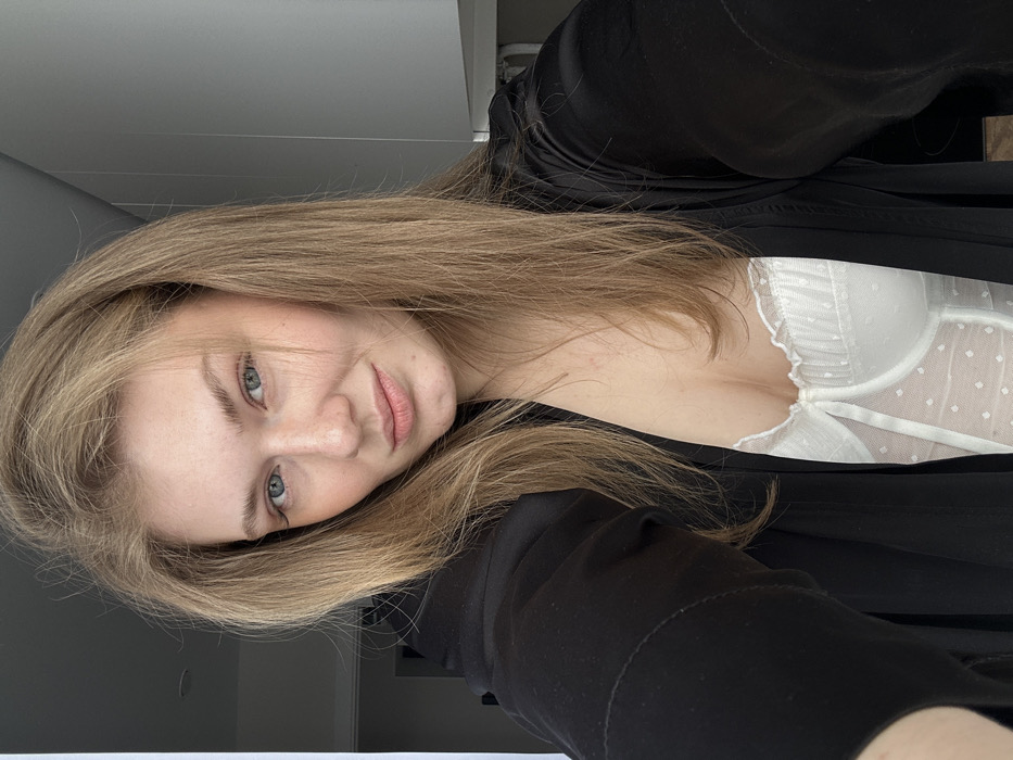 A single adult female is depicted in a medium-close-up self-portrait within an indoor setting. She has long, straight light brown hair, blue eyes, and light skin with visible freckles on her nose and cheeks. Her lips are slightly parted, and she gazes directly at the camera with a subtle smile. She wears an open, long-sleeved black garment, consistent with a robe or jacket, over a white structured undergarment. This white garment features a ruffled top edge and a sheer, polka-dotted section over the torso. Her left forearm is partially visible on the left side of the frame, indicating she is holding the camera. The background consists of light-colored, possibly white or light gray, walls. A light gray, rectangular object, possibly a cabinet door, is visible in the upper right quadrant. A darker area, resembling an open doorway or corridor, is present on the far left. The specific location within the city of Kazan, Russia, is not discernible from the visual information provided in the image.