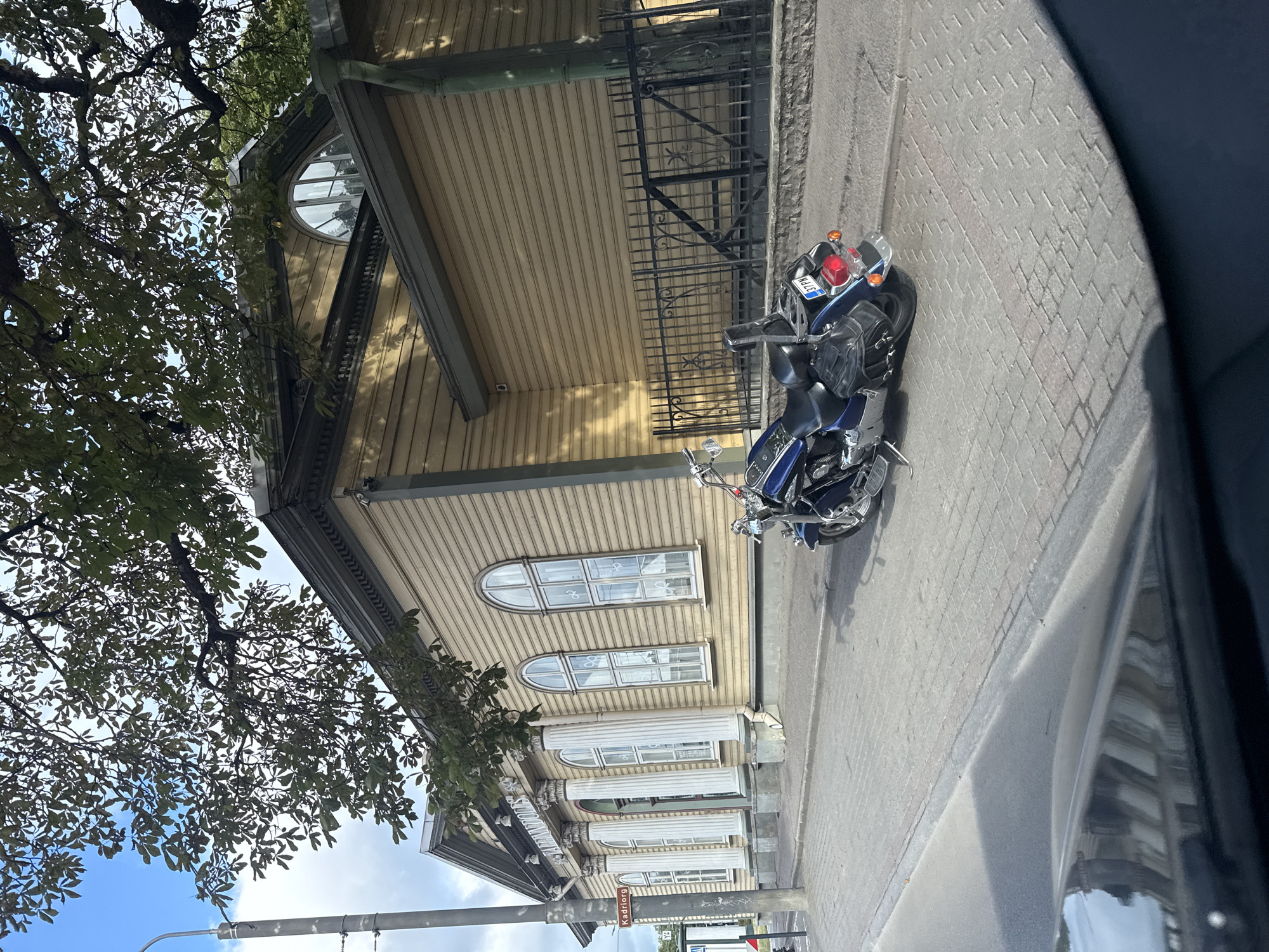 A blue motorcycle is parked on a cobblestone sidewalk in front of a historic yellow wooden building with white columns and arched windows. The building has a sign above the columns that reads "STADIONDOODES". A street sign on a pole to the left of the building reads "Kadriorg". The street is paved with asphalt, and a tree with green leaves overhangs the scene. The sky is partly cloudy. The motorcycle has a license plate that reads "37PN". The image appears to have been taken from inside a vehicle.