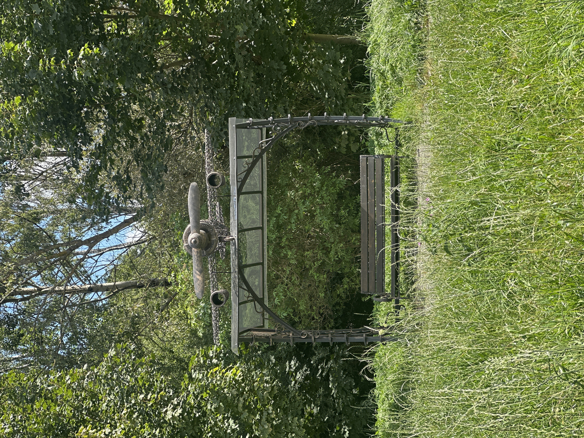 The image shows a metal swing set with a bench, adorned with an airplane propeller and engine parts on top. The swing set is located in a grassy area with trees in the background. The scene appears to be a park or outdoor recreational area. The location is identified as Minsk, Belarus. No people are present in the image.