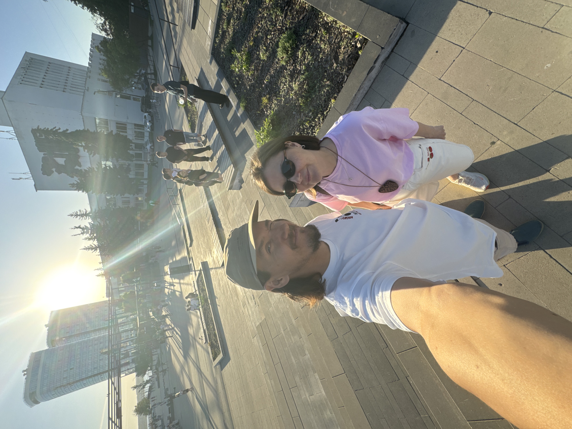 Two individuals are visible in the image, seemingly in the midst of taking a selfie. The man is wearing a white t-shirt and a hat, while the woman is wearing a pink t-shirt and sunglasses. The location appears to be a public space with paved stone. In the background, buildings are seen, and a group of people walking nearby. In the distance, the sun is shining brightly, creating a lens flare effect. The buildings in the background match the architecture of Izhevsk, Russia.