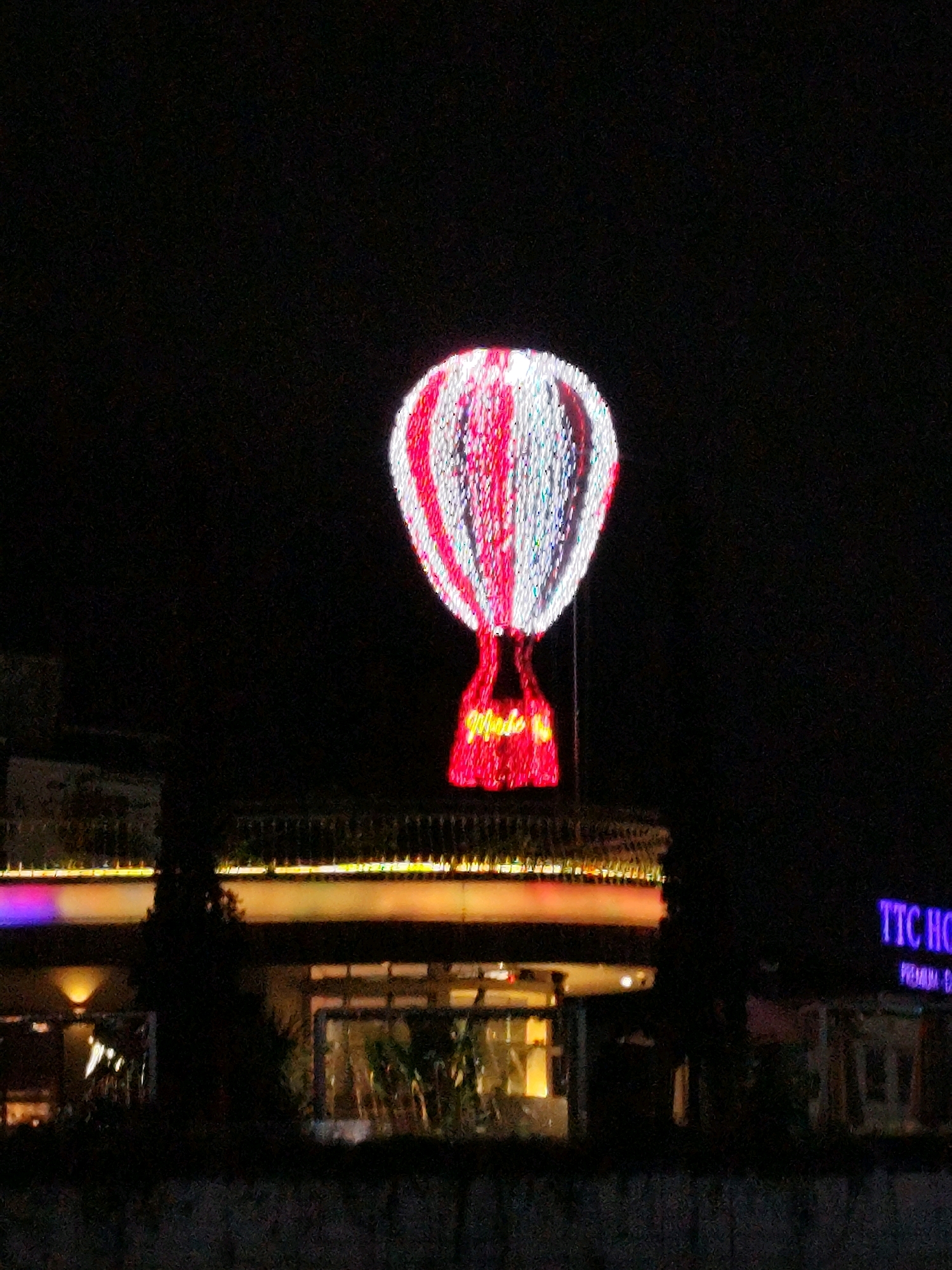 The image captures a nighttime view of a decorative hot air balloon illuminated by red and white lights, suspended above a building with a luminous facade. Below the hot air balloon, a sign that reads "Dalat" in cursive neon lights is visible. The building is lit with horizontal rows of yellow and purple lights. To the right of the building, a sign for "TTC Ho" with "PREMIUM" underneath is illuminated in purple and white. The scene appears to be an outdoor night setting, possibly a tourist attraction or a decorated building in the city of Da Lat, Vietnam, known for its unique architecture and festive light displays. The foreground shows a dark, blurred area that could be a fence or a hedge, with the lighting from the building reflecting on it.