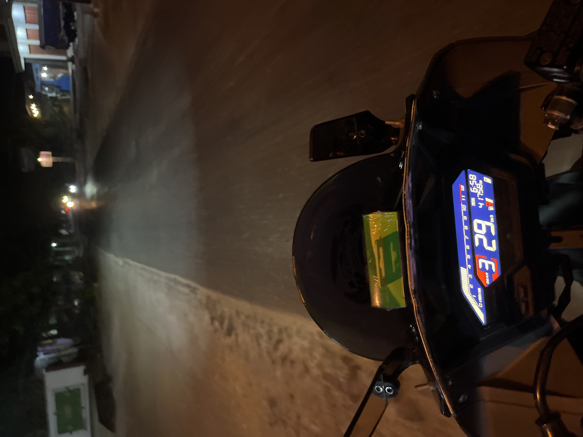 The image is taken from the perspective of someone riding a motorcycle at night on a road in Pecangaan, Indonesia. The foreground features the motorcycle's digital dashboard, displaying a gear of "3", a speed of "29 km/h", a total distance of "41758" and a time of "6:58". A green and yellow package is affixed to the windshield. The road ahead is visible, illuminated by the motorcycle's headlight and streetlights. On either side of the road are buildings and trees, characteristic of a semi-urban or rural area. The overall scene depicts nocturnal travel through a sparsely lit environment.