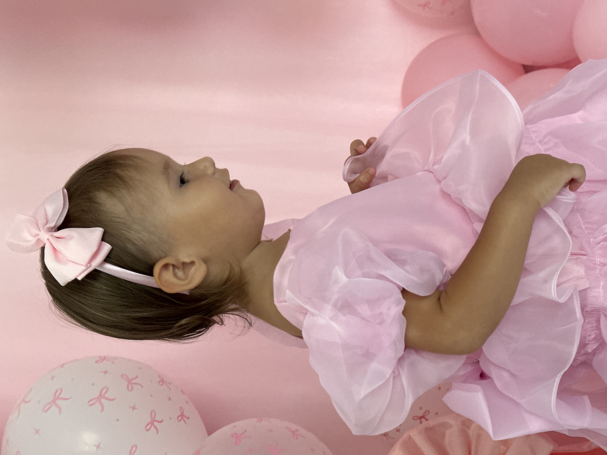 A young child is lying on a pink fabric backdrop, surrounded by pink balloons. The child is looking up and to the right, with their head tilted back. They are wearing a pink dress with ruffled sleeves and a pink bow in their hair. A pink balloon with a bow pattern is visible in the lower left corner of the frame. The scene appears to be a birthday celebration or a photoshoot.