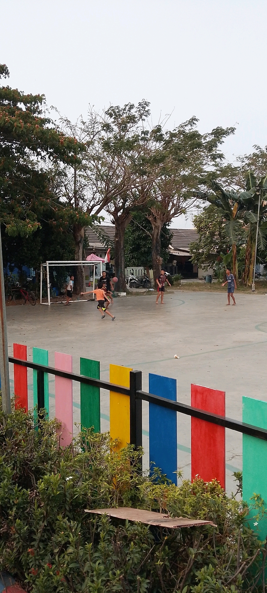 In Satriamekar, Indonesia, on a concrete court, a group of children are playing soccer. A white soccer goal is visible on the left side of the image. The court is surrounded by a fence with colorful vertical panels in red, pink, green, yellow, blue, and green. Trees with bare branches and some with green leaves are present behind the court. A few buildings and a parked motorcycle are also visible in the background. The sky is overcast.