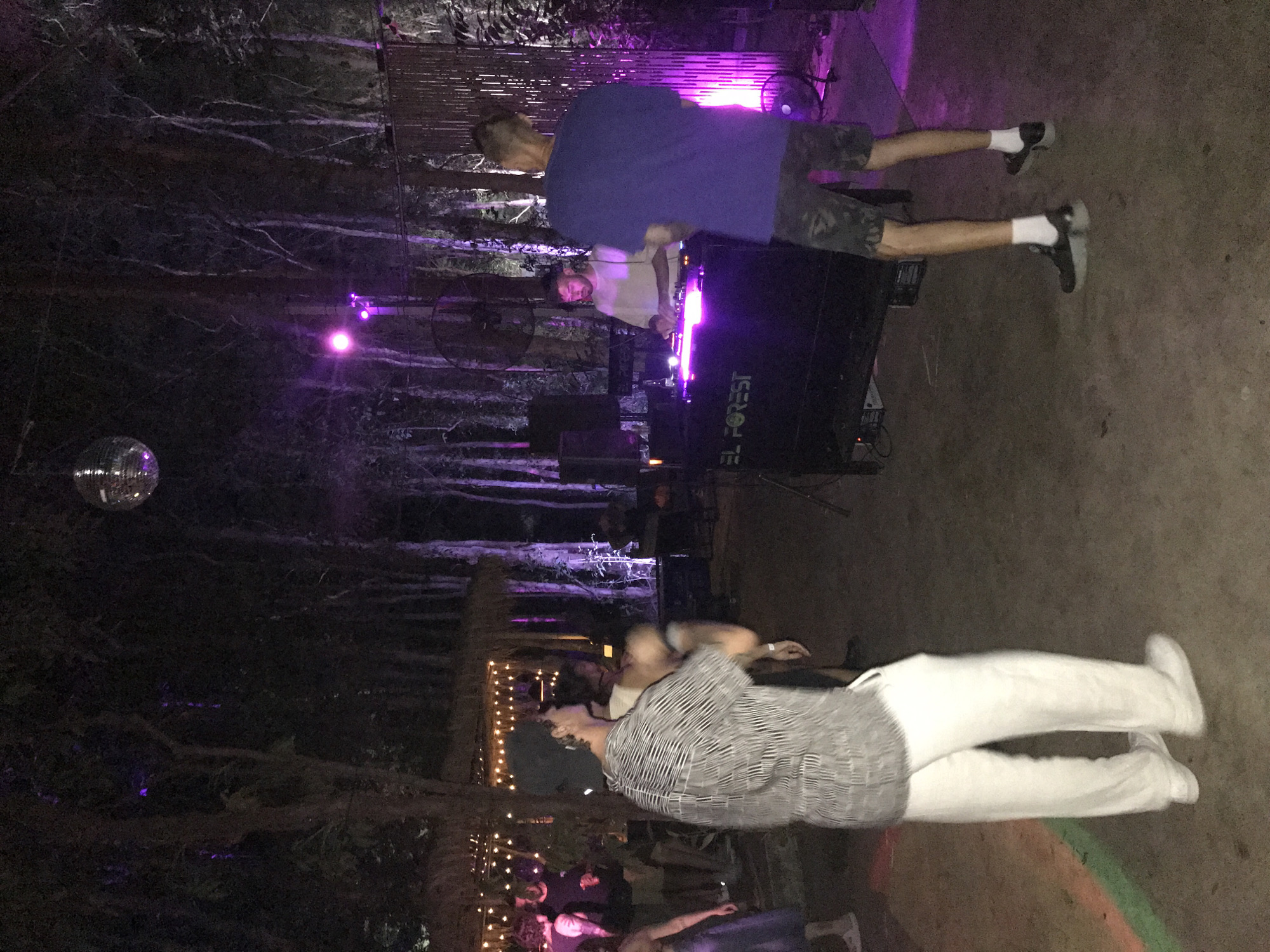Scene at night in Baan Mae Nam, Thailand, with a DJ setup labeled "EL FOREST" bathed in purple lighting. A disco ball hangs from above. Two men stand near the DJ equipment; one wears a blue shirt and camouflage shorts, and the other wears a white t-shirt. Another person in white pants and a striped shirt is in the foreground. Additional people are visible in the background near string lights. Trees surround the area.