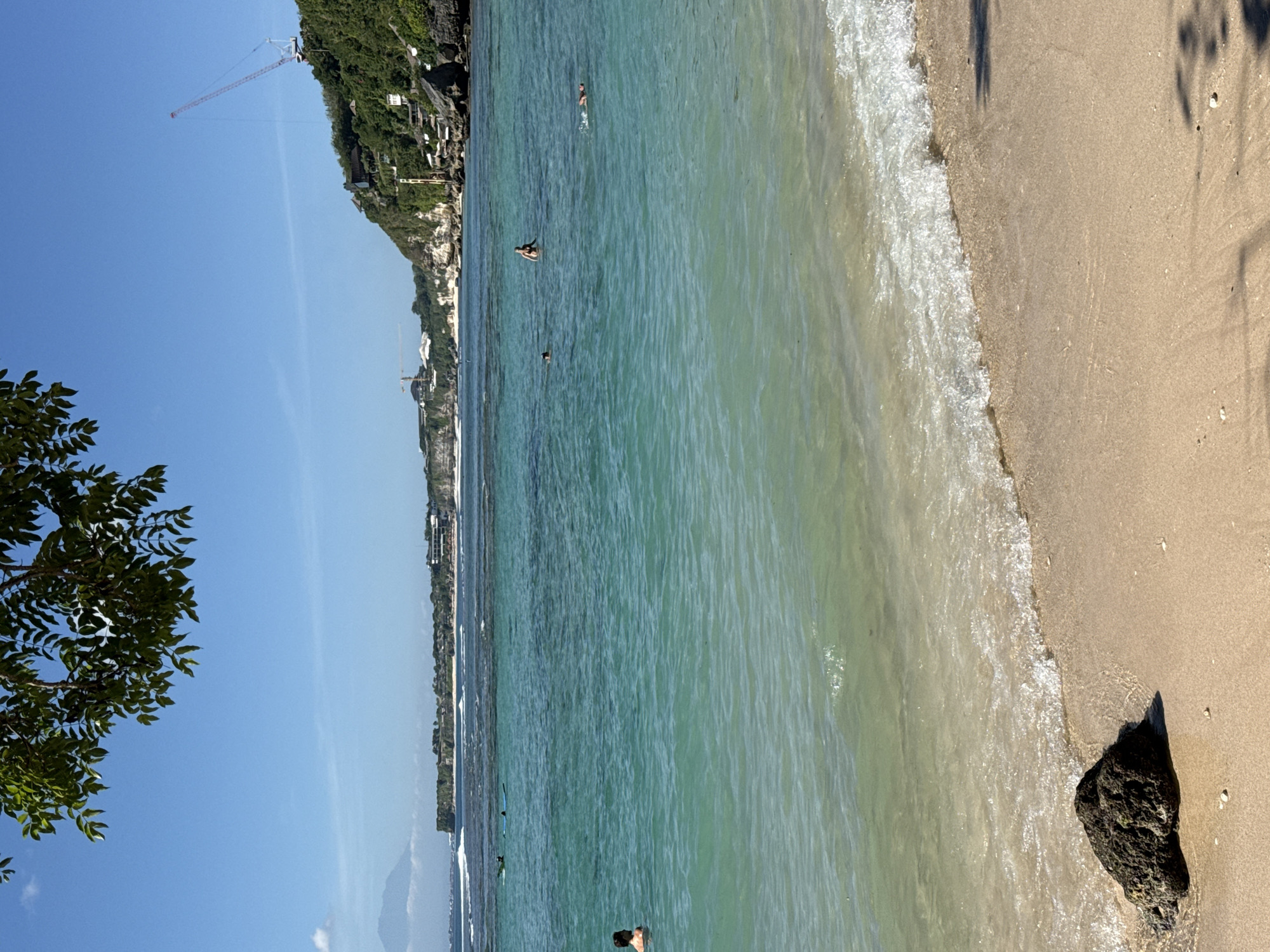 The image depicts a sunny beach scene in Kuta Selatan, Indonesia. The shoreline features light brown sand meeting turquoise water with gentle waves. Several people are swimming in the water, and a few small white shells are scattered on the sand. A dark rock sits on the beach near the water's edge. In the background, a cliff with lush vegetation and buildings runs along the coast. A construction crane is visible against the clear blue sky in the far distance. A tree with green leaves is in the foreground. The shadows suggest the photo was taken in the morning or late afternoon.
