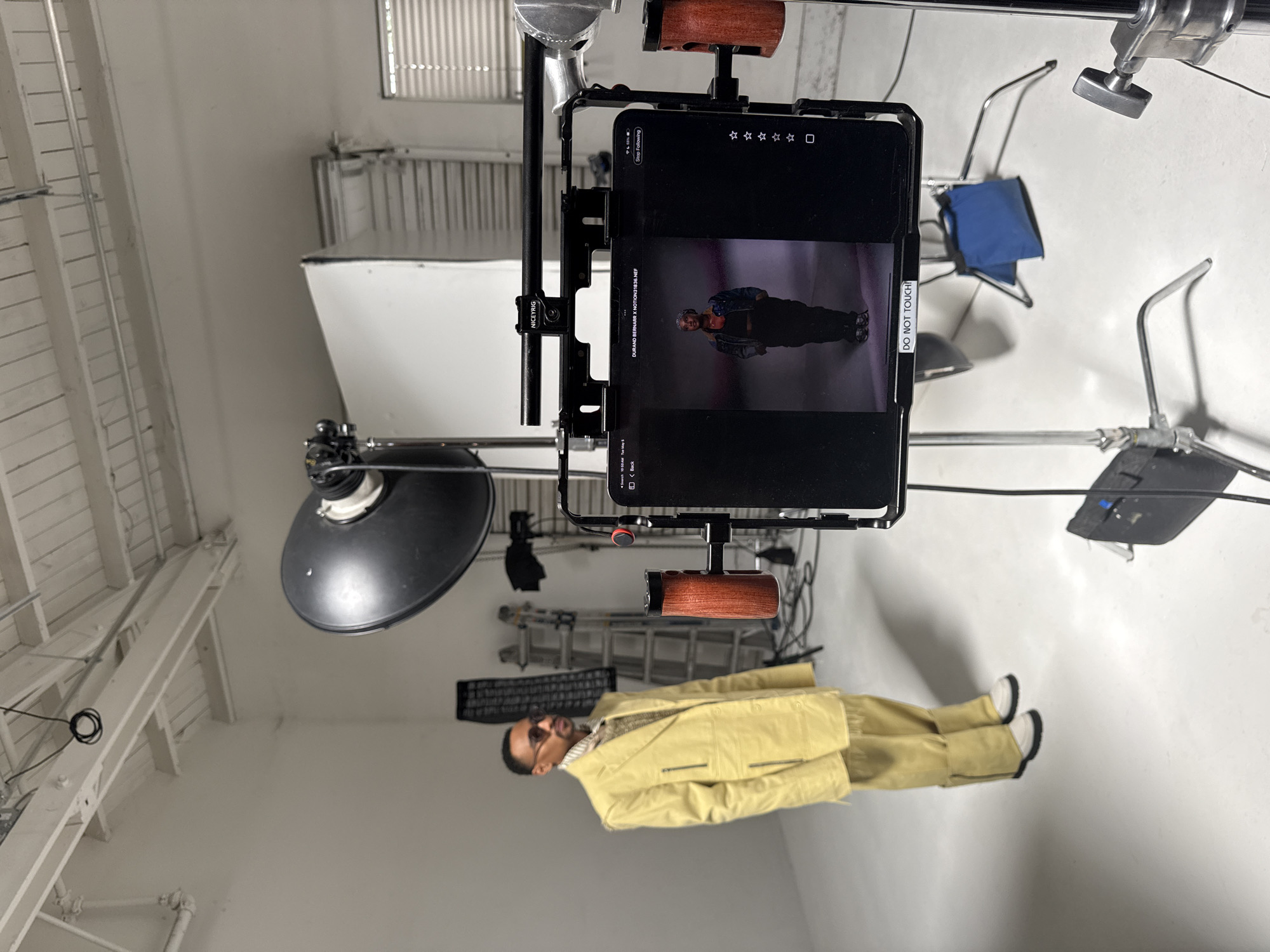 The image captures a scene inside a studio, likely for a photo or video shoot in Los Angeles, United States of America. A man dressed in a light yellow suit is lying horizontally on what seems to be a white floor. A large black reflector light fixture hangs from the ceiling, and a white backdrop is visible. The image also features a tablet, possibly an iPad, mounted on a metal rig, displaying a screen showing a picture of the same man. The focus is on the technical setup, as evidenced by the various stands, clamps, and riggings in the frame. The environment is well-lit, emphasizing the studio setting.