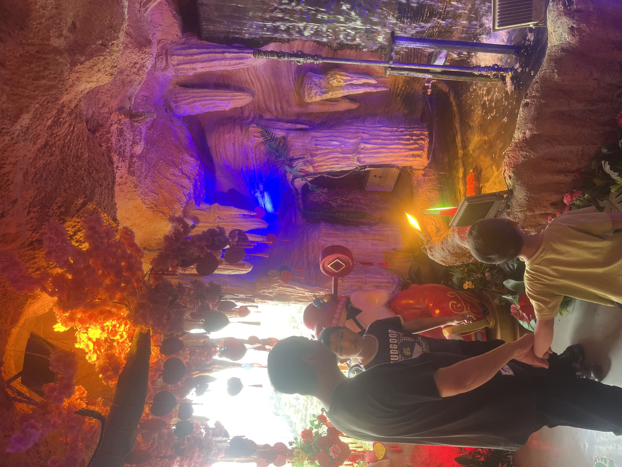 This vertical image depicts an elaborately decorated artificial cave interior, likely a themed attraction in Yuzhong District, China. The scene is awash in dramatic, multi-colored artificial lighting, creating a fantastical and surreal atmosphere.
The cave walls are intricately sculpted to resemble natural rock formations, featuring textures and stratified layers in shades of brown, pink, and beige. These surfaces are illuminated by a dynamic array of lights: a prominent blue glow emanates from one section, while purple and pink lights accent other areas. Warm orange and yellow light brightens the left side, particularly highlighting numerous artificial cherry blossom trees with their luminous pink and orange blooms. Red lighting is also visible near the ground level.
A waterfall feature is visible at the top of the cave, with water flowing over textured surfaces and metal structures. Various artificial green plants, including ferns and other foliage, are strategically placed throughout the rockwork. Decorative elements hang from the ceiling, including red circular lanterns and flat, heart-shaped ornaments. A larger, brightly colored red sculpture or object is situated near the center of the scene.
Four individuals are present, visible in the foreground and towards the right. One adult, seen from behind wearing a black t-shirt with the text "UDGOOD" clearly visible on the back, is holding hands with a younger child dressed in a lighter-colored shirt. Another adult and child are partially visible further back. They appear to be walking through or observing the unique environment. A bright, overexposed light source or window is present on the far left, partially silhouetting some of the figures.
The overall impression is that of a modern, immersive indoor leisure or entertainment space designed to captivate visitors with its blend of simulated natural features and vibrant, artificial illumination.