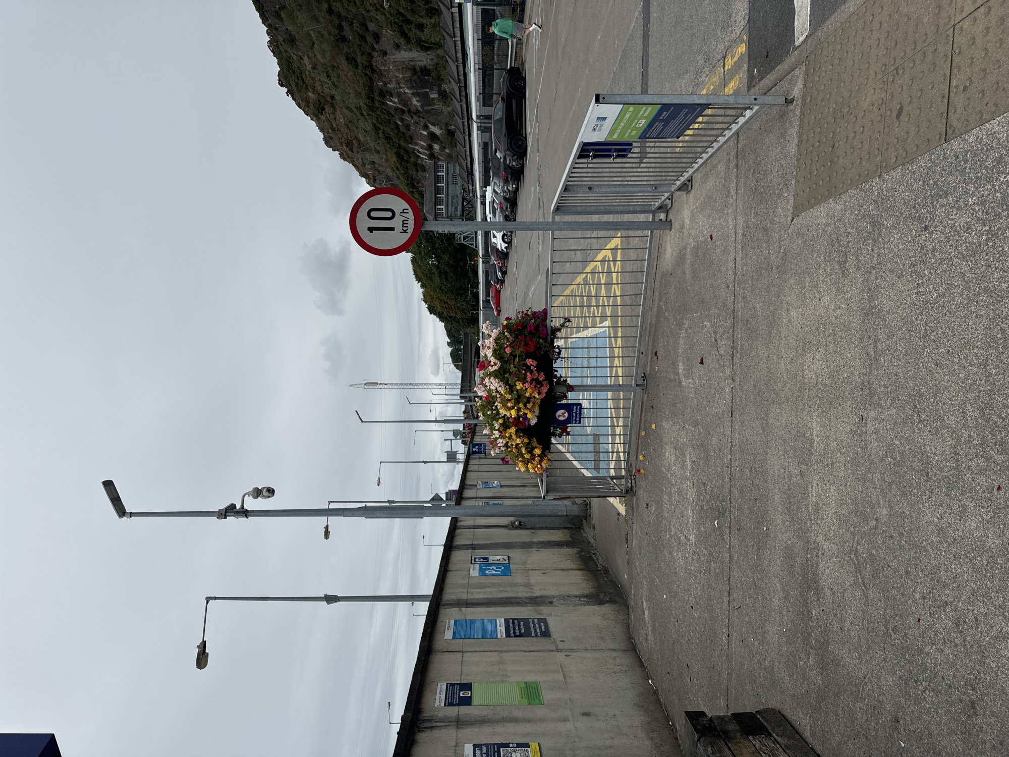 The image depicts an outdoor parking lot scene under an overcast sky. In the foreground, a concrete walkway leads towards a metal barrier, beyond which is a parking area marked with yellow lines. A flower basket, overflowing with colorful blooms, hangs from a post near the barrier. A circular speed limit sign indicating "10 km/h" is mounted on a pole in front of the parking lot.
The parking lot contains several parked cars, including a black SUV, a white SUV, and a red car. In the background, a railway station with a platform and a train are visible. A steep, vegetation-covered hillside rises behind the station. A person in a green hoodie and light shorts is walking away from the camera in the parking lot. The wall on the left side of the image has several posters and parking signs attached to it. The overall environment suggests a public transportation hub or a facility with a speed restriction. The location is identified as Unknown, Ireland, with no further specific details available from the image itself.