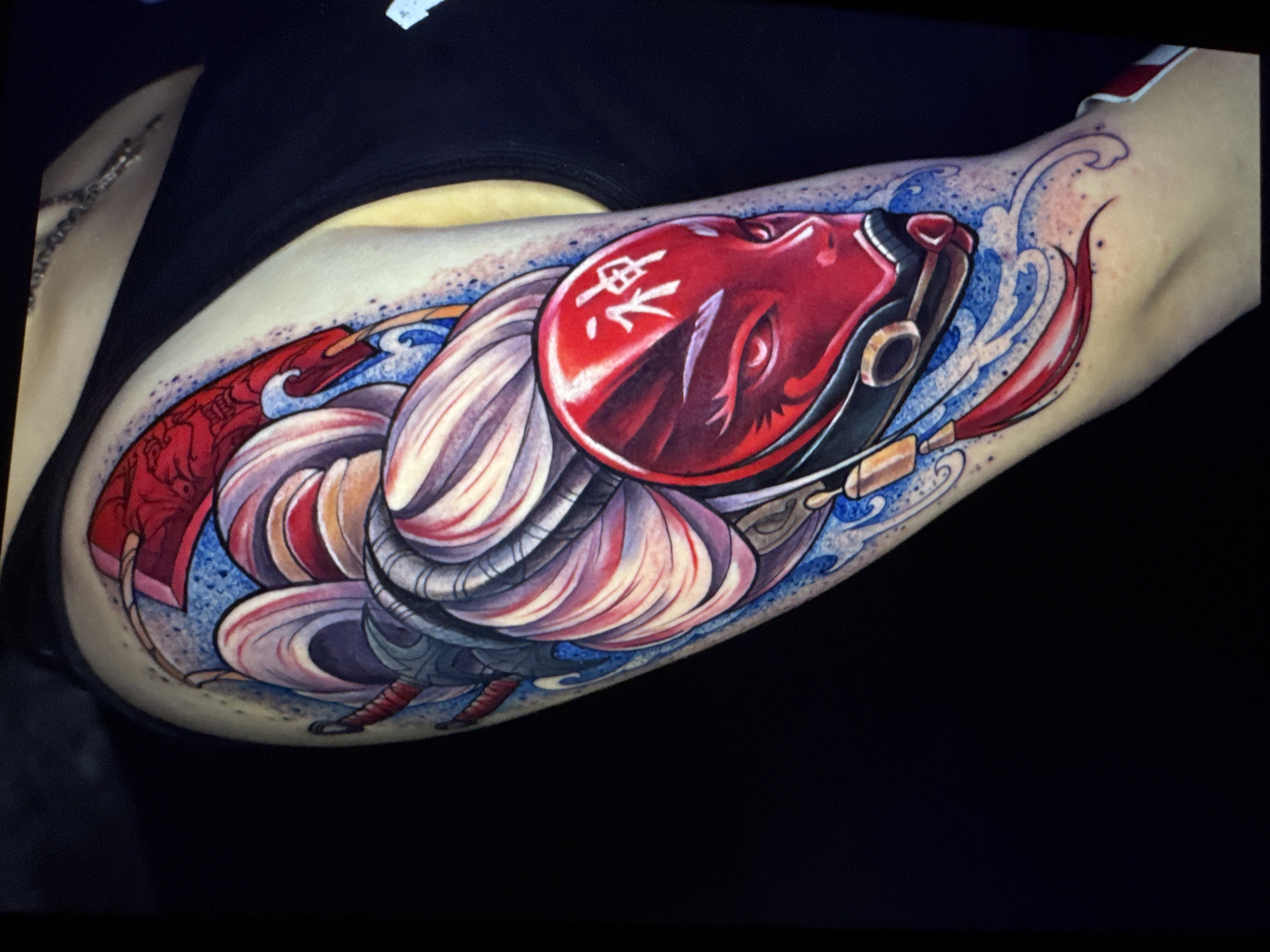 The image shows a close-up of a tattoo on a person's arm. The tattoo depicts a stylized, red fish with flowing fins and intricate details. The fish appears to be swimming in water, represented by blue wave-like patterns and speckles of blue and red ink. The fish has a prominent red head with a black outline, and a white symbol or characters are visible on its forehead. The fins are rendered in shades of white, pink, and purple, creating a sense of movement and fluidity. The overall style of the tattoo is vibrant and detailed, with strong use of color and shading. The background is dark and plain, emphasizing the tattoo itself. The context of the image is a person displaying their tattoo, likely for artistic or personal reasons.