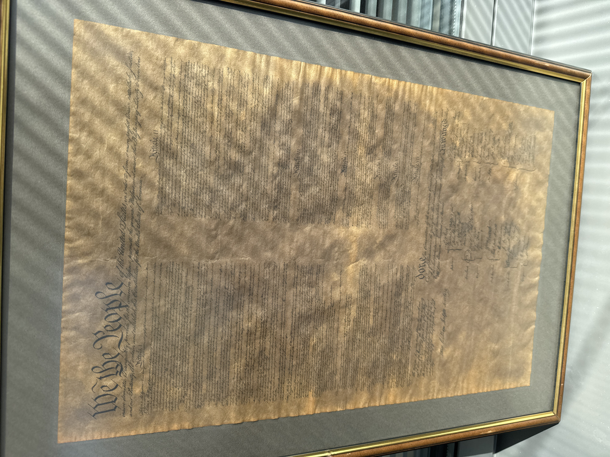 The image shows a framed document, specifically a replica of the United States Constitution, as evidenced by the large text "We the People" prominently displayed on the left. The document is aged, featuring a yellowish-brown hue, and shows signs of wear and tear, possibly intended to mimic the original's appearance. It's framed with a brown and gold-colored border and appears to be mounted on a gray backdrop. The surrounding environment is bright, with visible light streaks suggesting that the photo was taken indoors near a window. There is no information in the image to suggest it is in Sofia, Bulgaria.