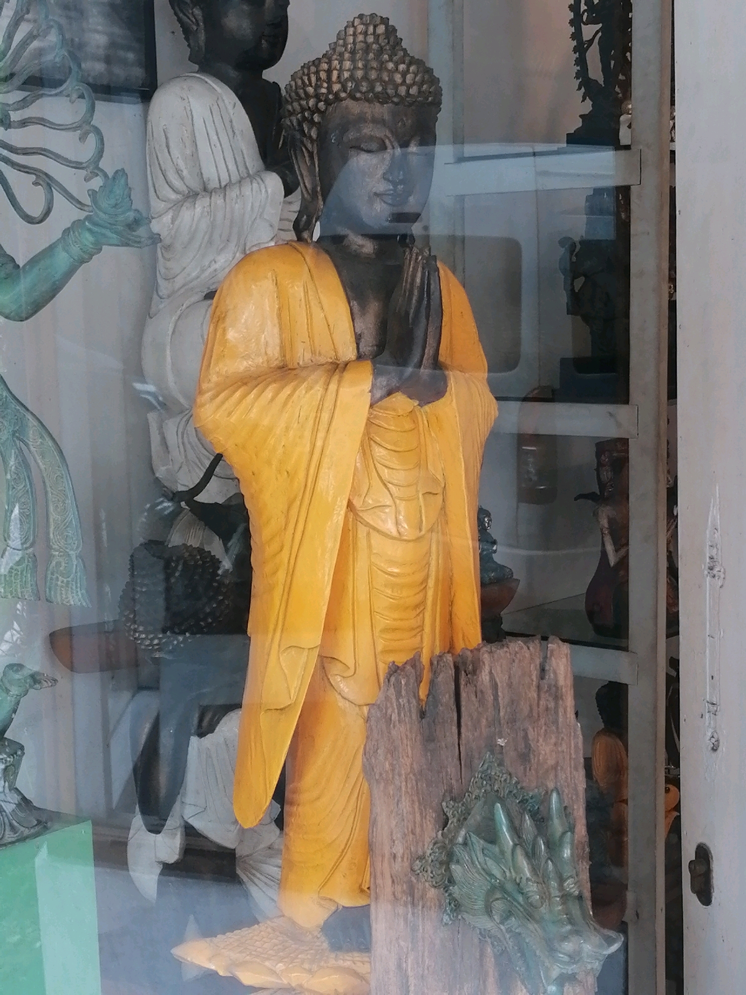 The image shows a display of religious statues, possibly in a shop or temple, in Ubud District, Indonesia. A large statue of Buddha, wearing a yellow robe and with hands pressed together in a gesture of prayer or greeting, dominates the center of the frame. The Buddha's head is detailed with curly hair and a serene expression. To the left of the central Buddha, another statue of Buddha, this one white and draped, is partially visible. Behind the central Buddha, other statues and religious artifacts can be seen on shelves, including what appears to be a dark-skinned statue of Buddha and a dark silhouette of a figure in a dynamic pose. In the foreground, a piece of weathered wood with an ornate, possibly dragon-like, metallic applique is propped up. Reflections on a glass surface, suggesting a display case or window, are visible throughout the image. The lighting appears to be indoor, with some natural light filtering through.