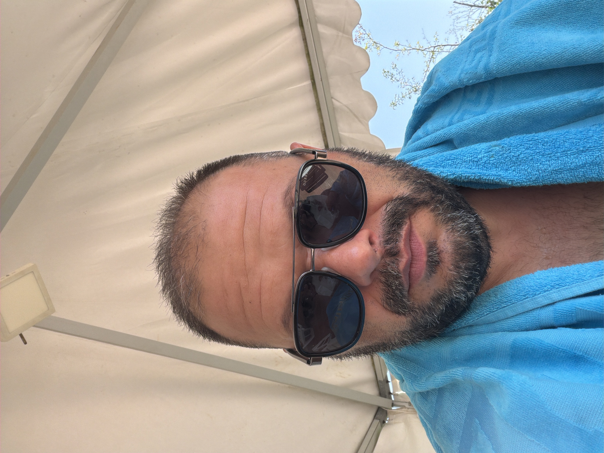 A man with a beard and sunglasses is pictured from the chest up, wearing a blue towel around his shoulders. He is outdoors, with a white canopy above him and a blue sky visible in the background. The image appears to be taken in Simeonovo, Bulgaria, possibly at a resort or outdoor recreational area.