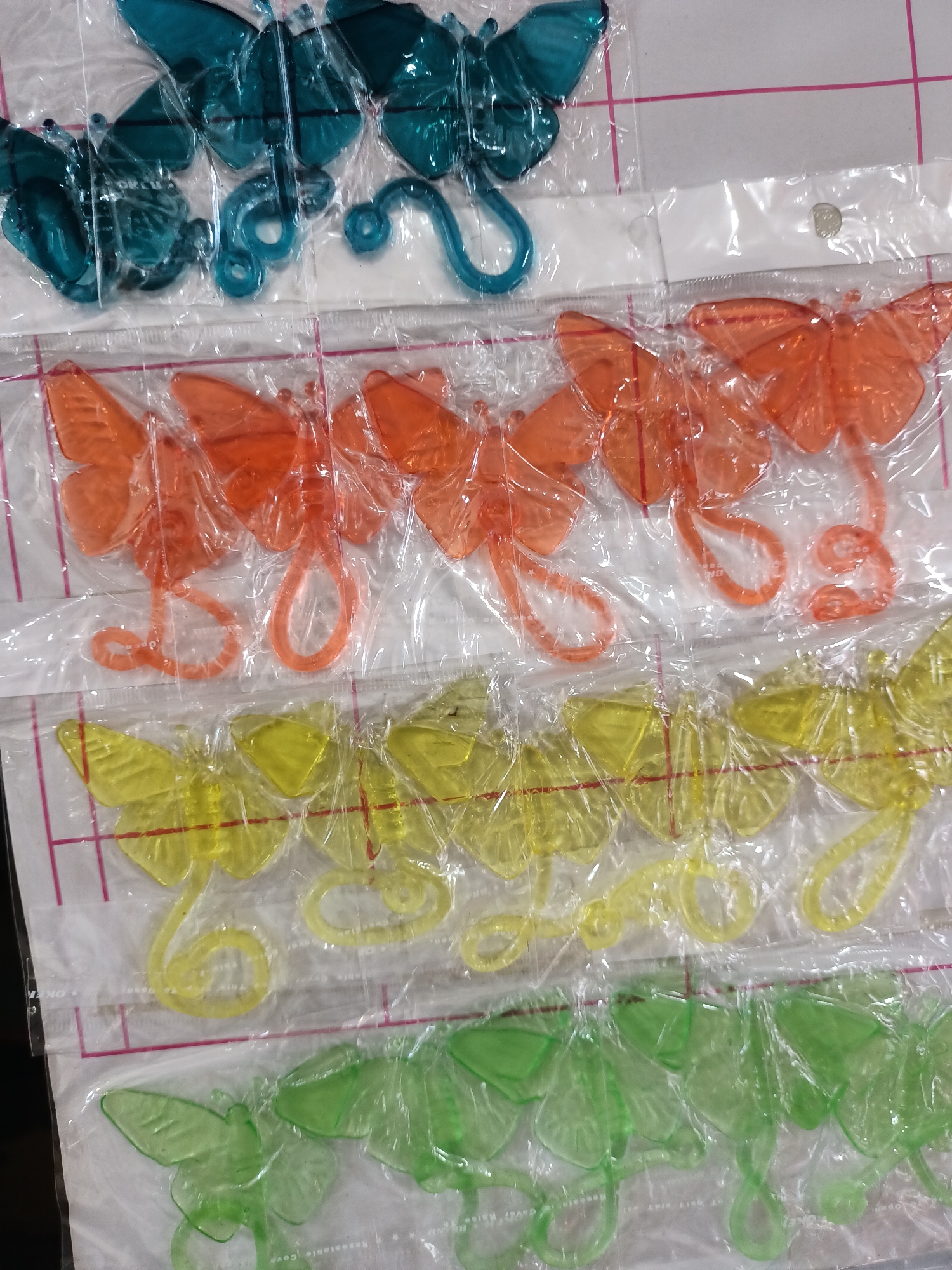 The image shows four rows of plastic butterfly-shaped ornaments, each row a different color: teal, orange, yellow, and green. The ornaments are individually sealed in clear plastic packaging. The background appears to be a white surface with a faint grid pattern, possibly a tabletop or counter. There are no people or any contextual clues to indicate a specific location in the city of Abay, Kazakhstan.