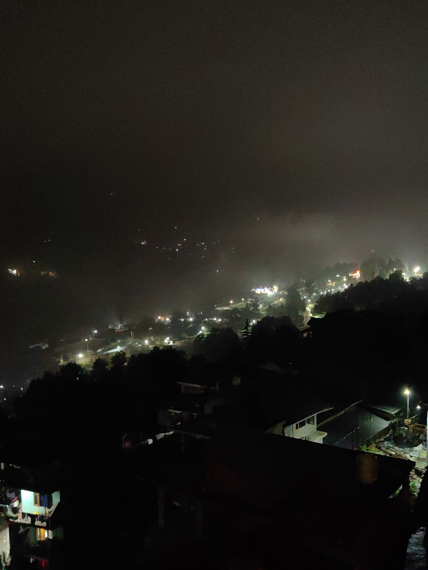 This is a nighttime, high-angle, medium-shot photograph of a dimly lit, mountainous town, likely Vashisht, India, with a hazy, foggy atmosphere. The foreground is dominated by dark, silhouetted structures and trees, obscuring much of the detail. In the mid-ground and background, the town is illuminated by numerous scattered lights, suggesting buildings and streetlights nestled into the undulating terrain. Some of these lights appear to be on pathways or roads winding through the landscape. A faint mist or fog permeates the scene, softening the edges of distant lights and creating a diffused glow. The sky is dark and overcast, with no visible stars or moon. There are no discernible people or specific actions visible in the image, only the static presence of the illuminated settlement against the dark, misty backdrop. The overall impression is one of a quiet, diffusedly lit mountain village at night.