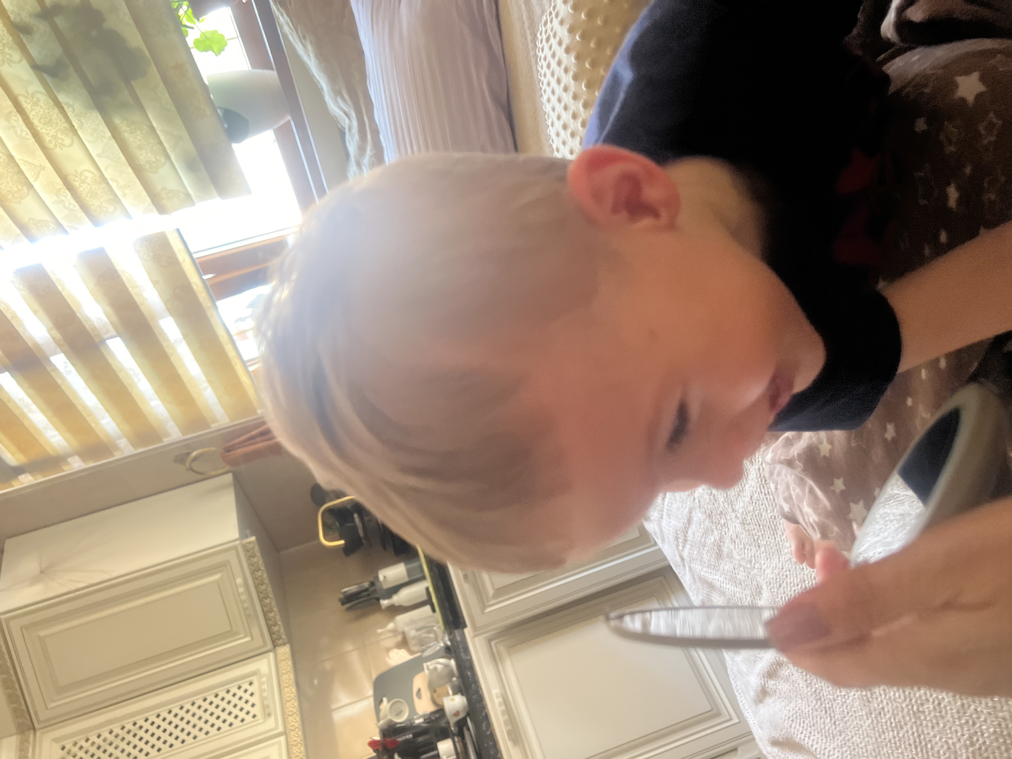 An indoor medium shot shows a fair-haired child in profile, looking at a mirror held by an unseen person's hand. The child is wearing a dark blue shirt, and their right cheek and ear are visible. They are lying on a textured blanket. In the background, kitchen cabinets with ornate detailing are visible on the left. A window with horizontal blinds is on the right, letting in bright sunlight. A plant is visible on the windowsill. The overall lighting is bright and diffused, with lens flare from the sun.