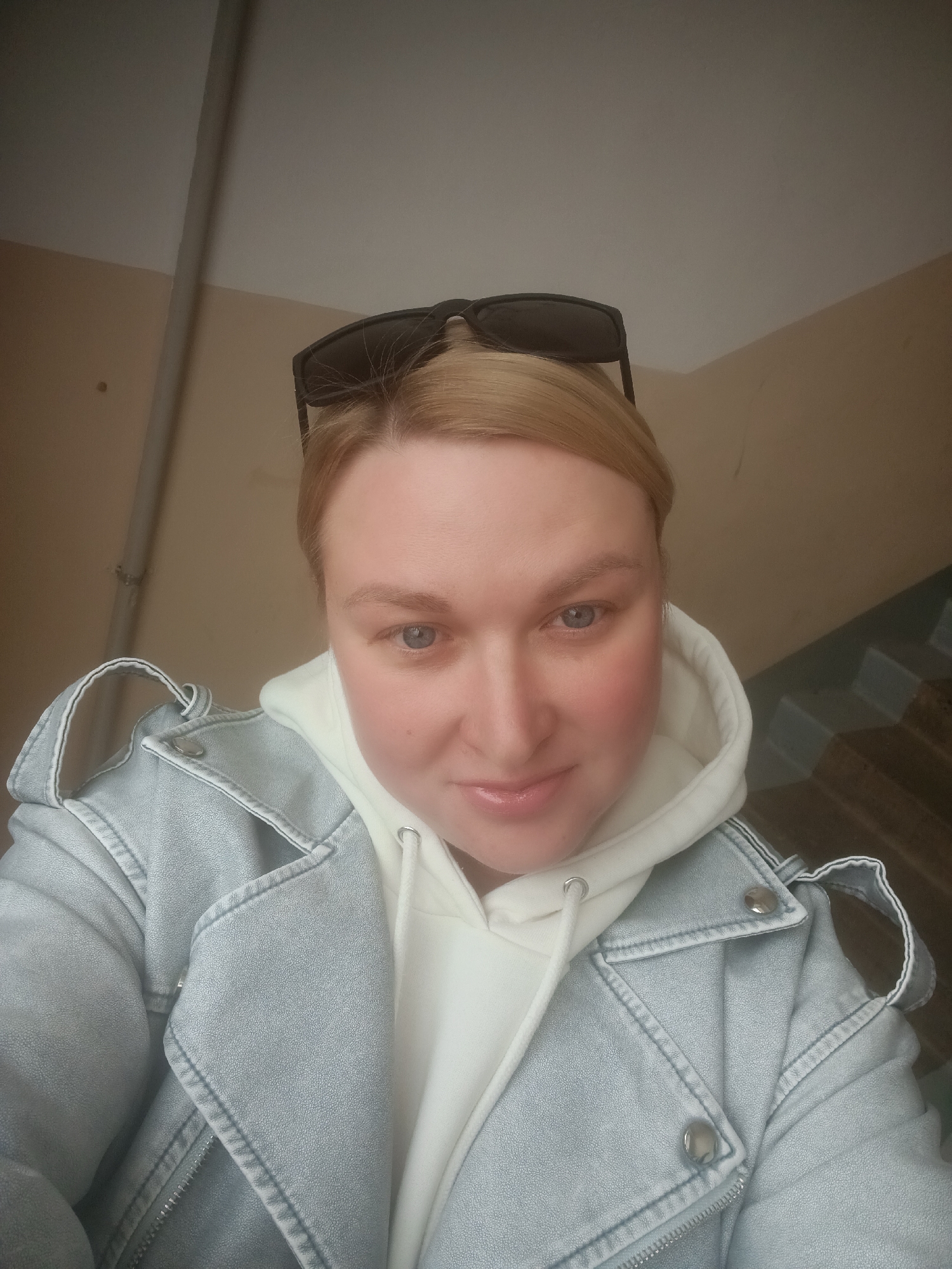 A selfie taken by a blonde woman in a light blue denim jacket over a white hoodie. She has sunglasses perched on top of her head. The background shows a wall with a light beige color and a stairwell with brown stairs. The location appears to be an indoor staircase within the city of Chaikovsky, Russia, based on the metadata.