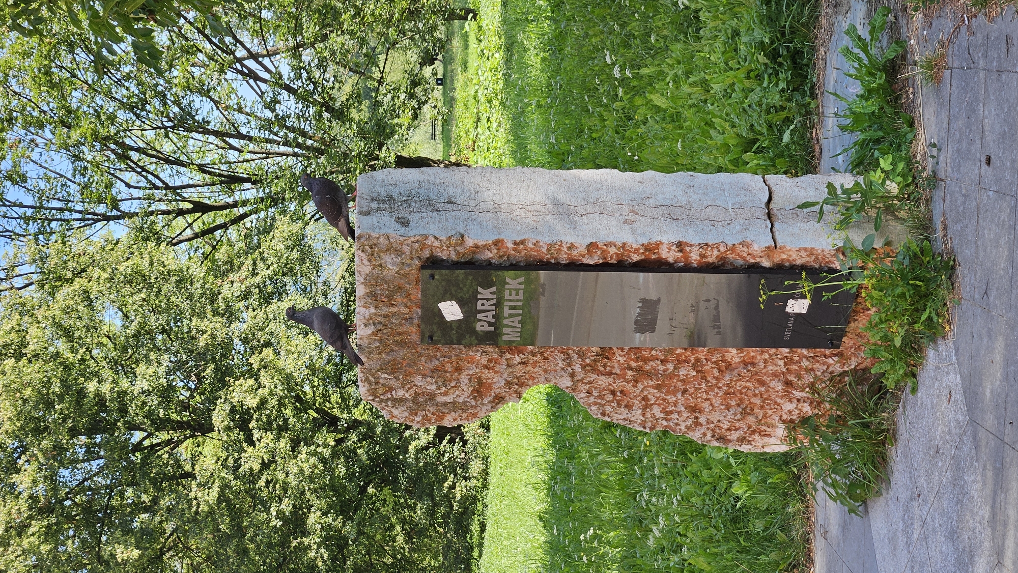 Two pigeons are perched on a stone monument in a park. The monument is roughly rectangular and appears to be made of two different types of stone, one reddish-orange and rough, the other grayish-blue and smoother. A black plaque is embedded in the monument, displaying the words "PARK MATIEK" in white lettering. Below this, faint text is visible, possibly a name or dedication. The monument stands in a grassy area with scattered white wildflowers. Trees with dense green foliage form a backdrop, and the sky is blue with some clouds. The ground in front of the monument is paved with gray concrete slabs. The park appears to be located in the city of Prešov, Slovakia.