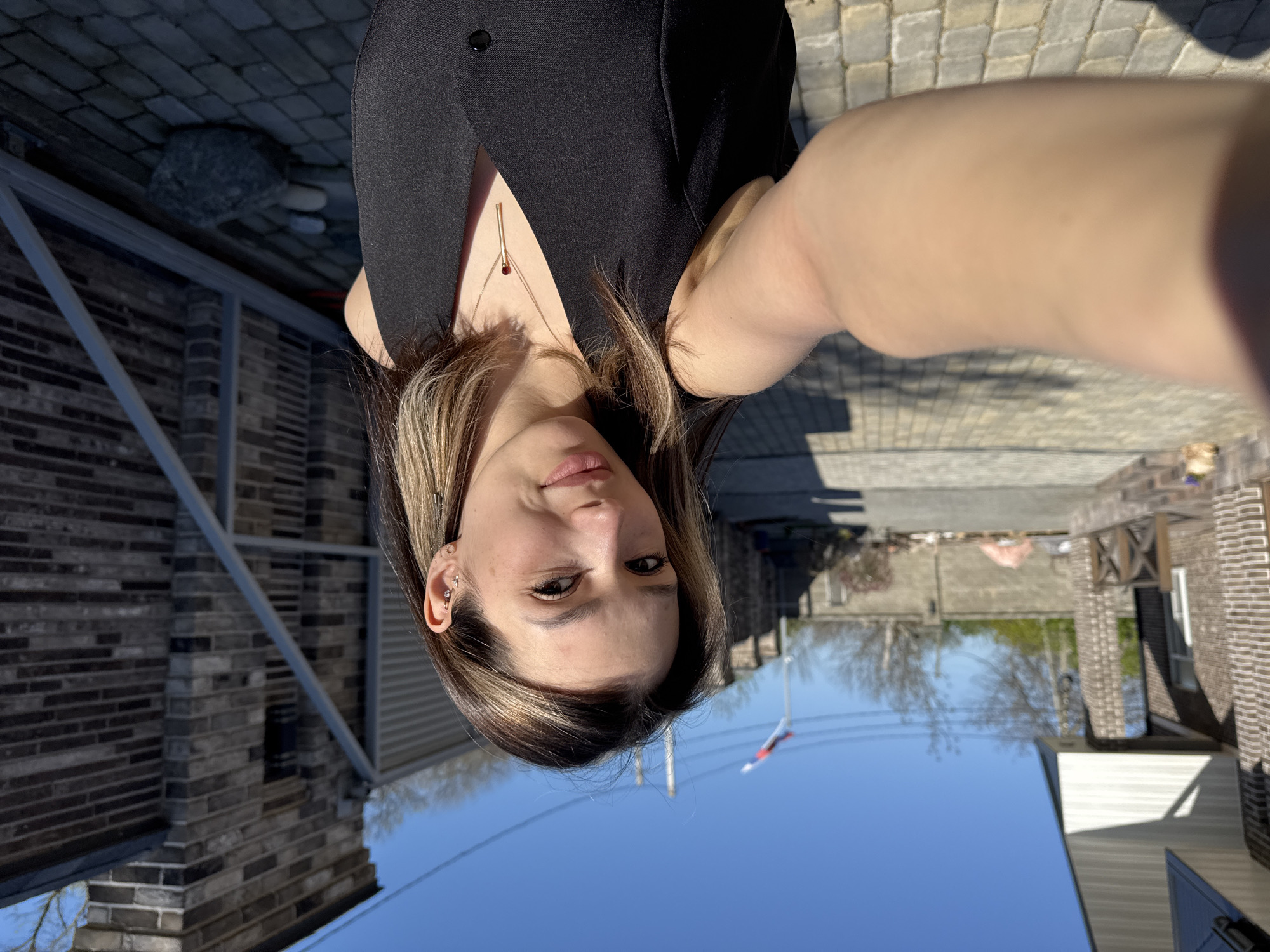 The image depicts a woman in a black top taking a selfie outside on a sunny day. She is wearing a necklace. Behind her is a scene that includes brick buildings and a clear blue sky. A flag is visible in the background. The ground is paved with square stones.