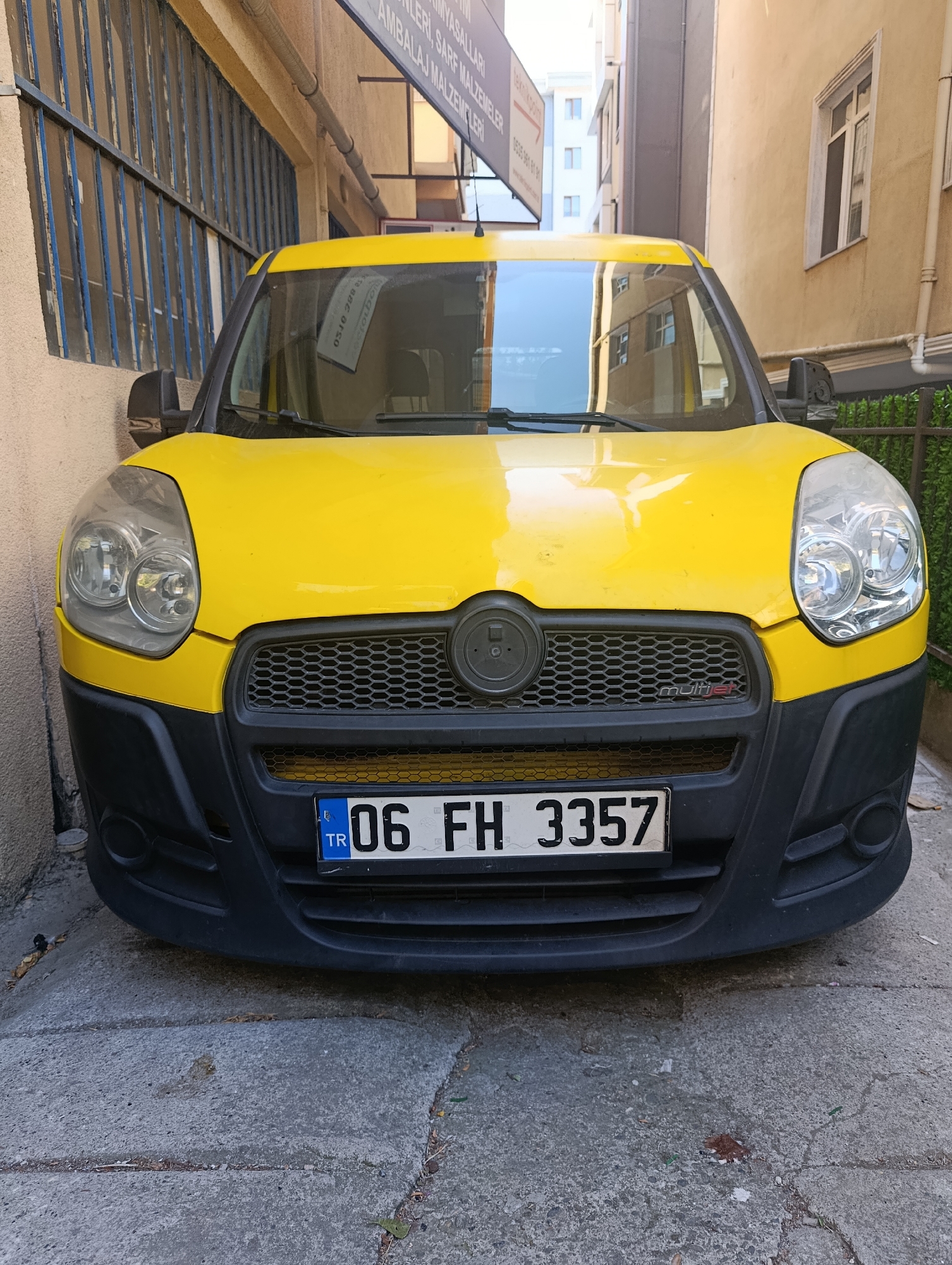 A yellow Fiat Doblo van is parked on a concrete path between two buildings in Ümraniye, Turkey. The van has a black bumper and a license plate that reads "06 FH 3557". The van's headlights are visible, and the windshield reflects the surrounding buildings. To the left of the van is a building with a blue-barred window, and to the right is a building with a cream-colored facade and a window. A metal fence with green foliage is visible behind the van. The ground is cracked and shows signs of wear and tear.