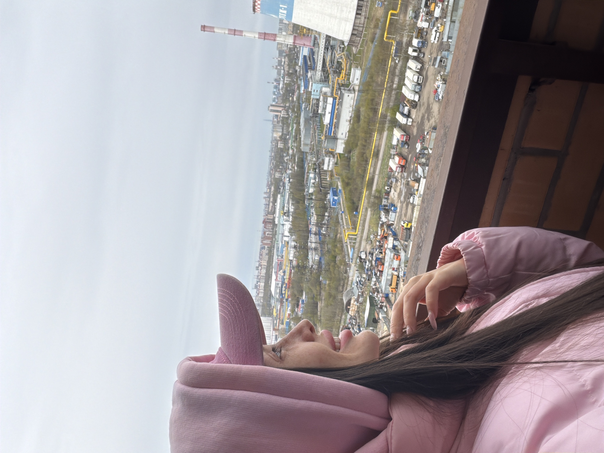 The image, oriented on its side, captures a woman observing an expansive industrial and urban landscape from a high vantage point, likely a balcony. In the foreground, a woman with long, dark hair, dressed in a pink hooded sweatshirt and a matching pink puffer jacket, is seen in profile. Her right hand, adorned with long, light-colored manicured nails, rests gently on her hair. She looks upwards and to the right, her expression appearing contemplative as she surveys the distant view.
Beyond a dark metal railing on the right, a sprawling cityscape unfolds under an overcast, daytime sky. The scene is dominated by industrial infrastructure, including numerous buildings that resemble factories or warehouses, and vast areas crisscrossed by yellow pipelines. A distinctive, tall industrial chimney, marked with red and white stripes and a blue section at its top, stands prominently in the mid-ground. The Cyrillic text "ТЭЦ-1" (TETs-1), which stands for "Thermal Power Plant-1," is clearly visible on the blue section of the chimney. Below, an extensive open area or parking lot is filled with a multitude of trucks and vehicles. The immediate right foreground also shows a section of a brick wall, confirming the elevated perspective. The industrial backdrop is consistent with areas found in cities like Saint Petersburg, Russia, where such power plants and industrial zones are common.
